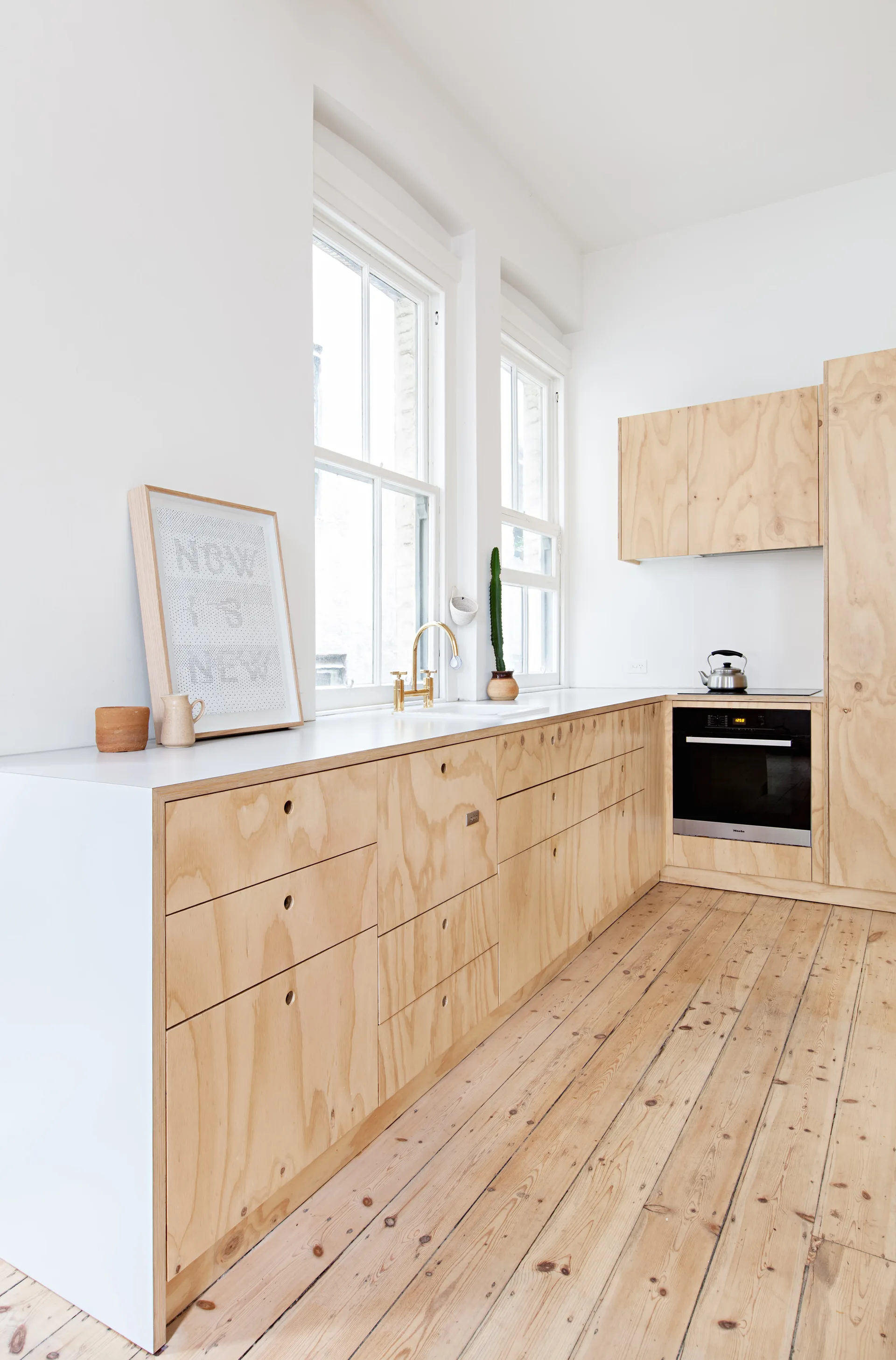 A bright kitchen features light-toned plywood cabinets with circular cutout handles, white countertops, tall windows, and natural wood flooring.