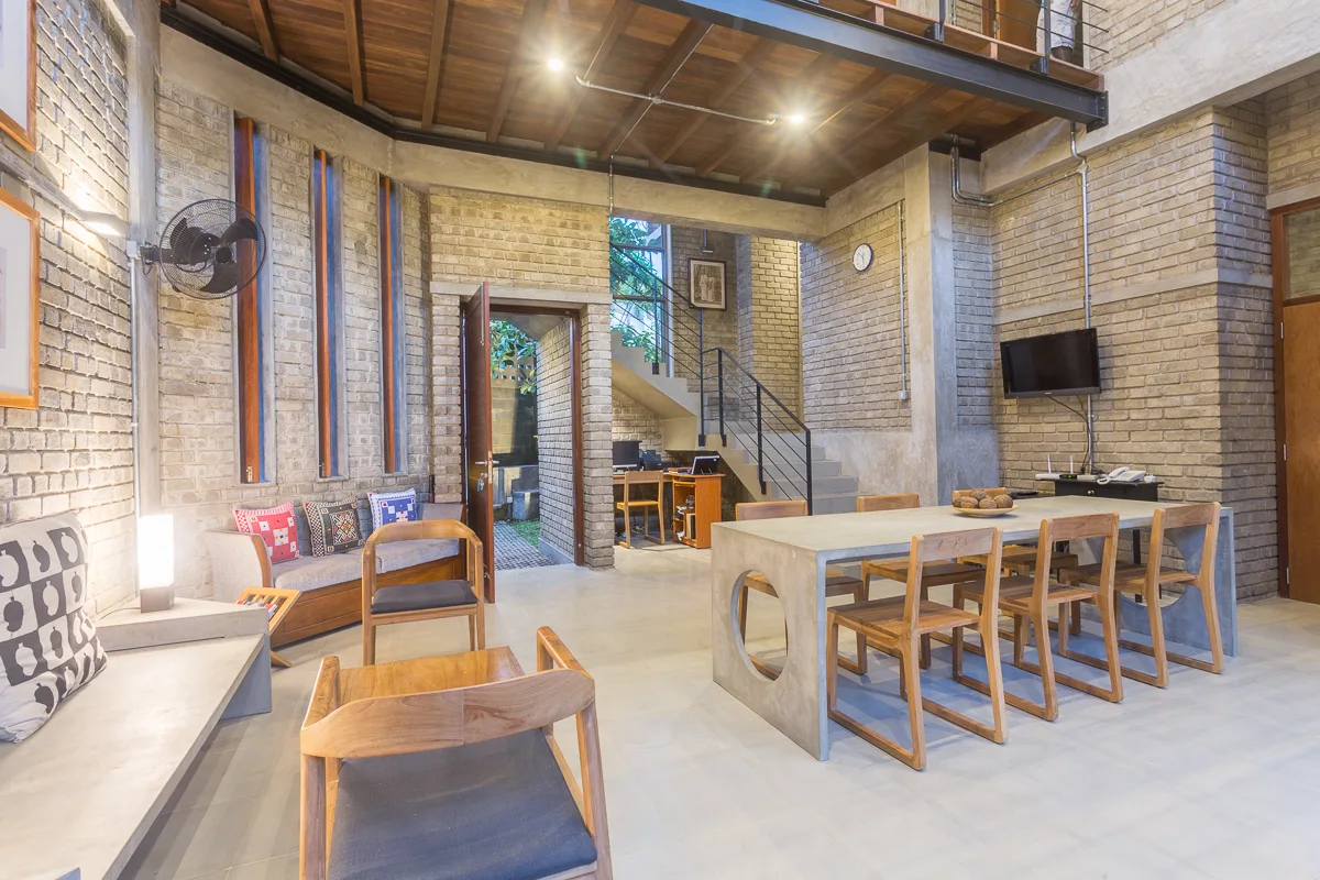 An industrial dining area with exposed brick walls, a long concrete table, wooden chairs, and a mezzanine level overhead.