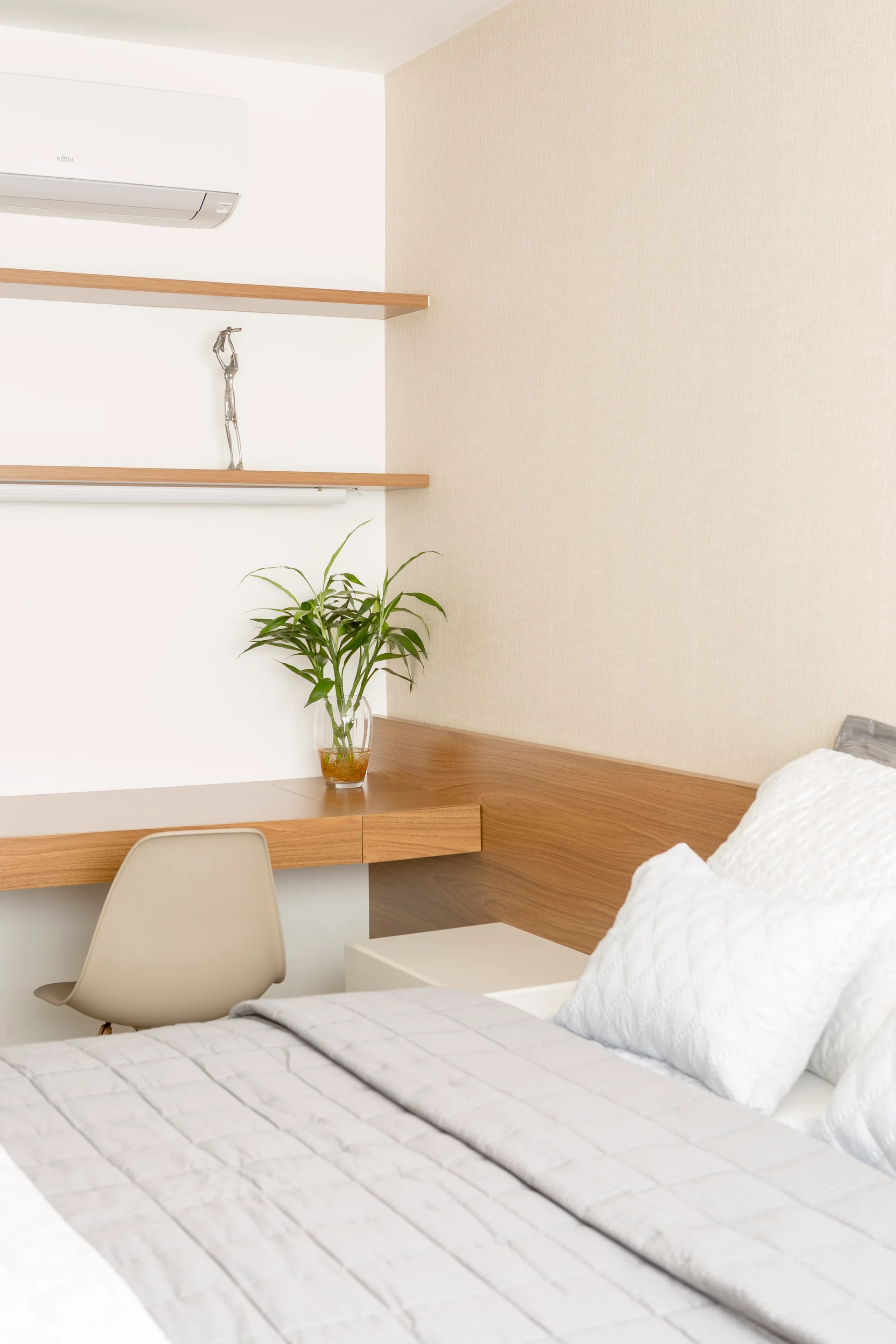 A modern bedroom features a light wood integrated desk and headboard, floating shelves, a beige chair, and grey quilted bedding.