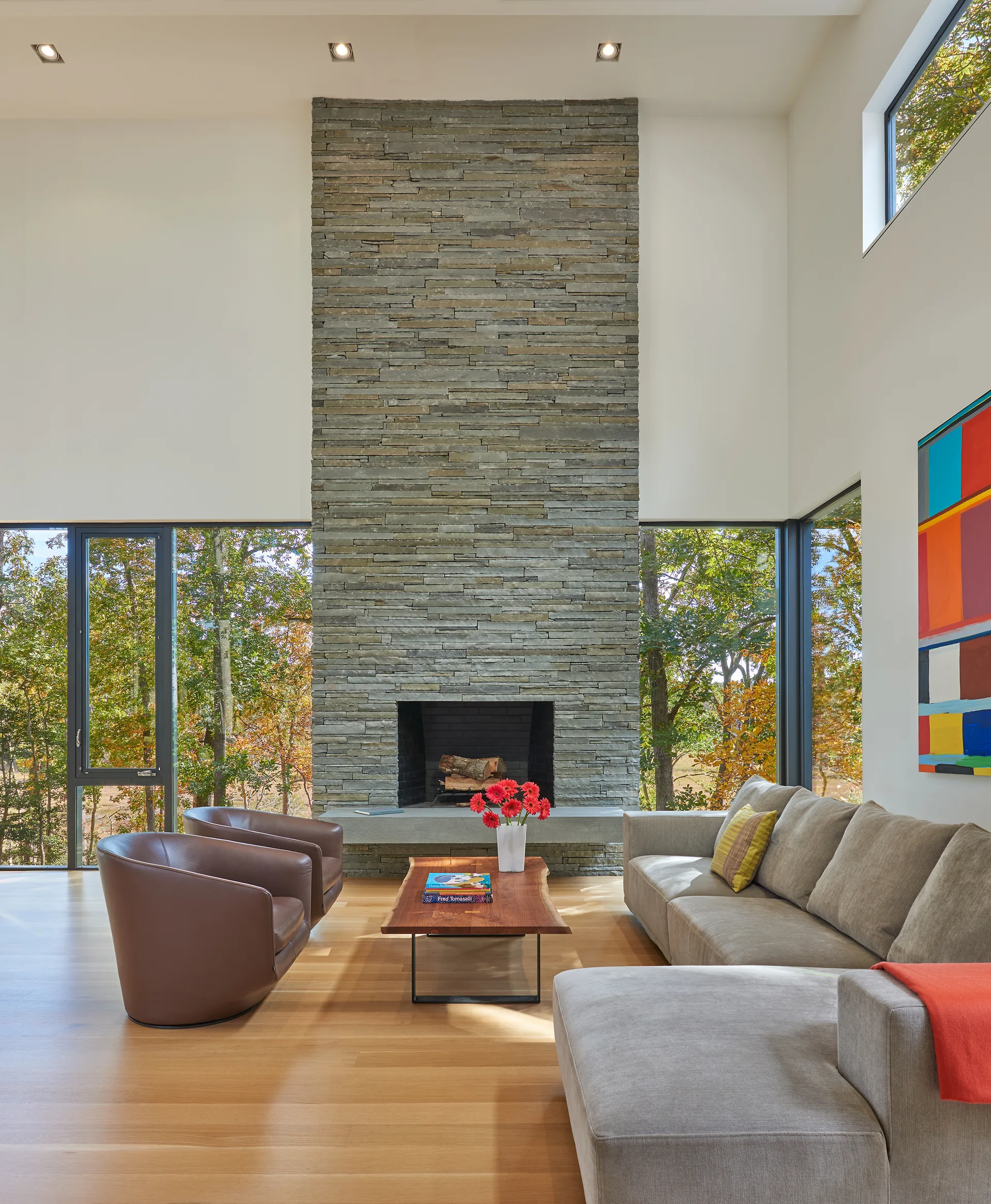 A bright living room features a tall gray stacked stone fireplace, a gray sectional sofa, two brown leather armchairs, and large windows.