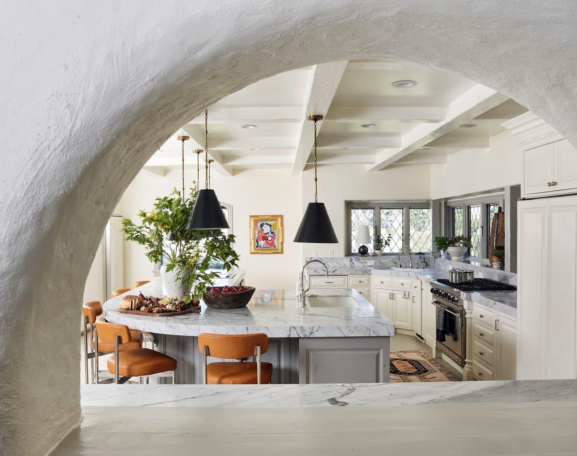 A kitchen with a marble-topped island and orange stools, framed by a stucco archway and illuminated by black pendant lights under a beamed ceiling.
