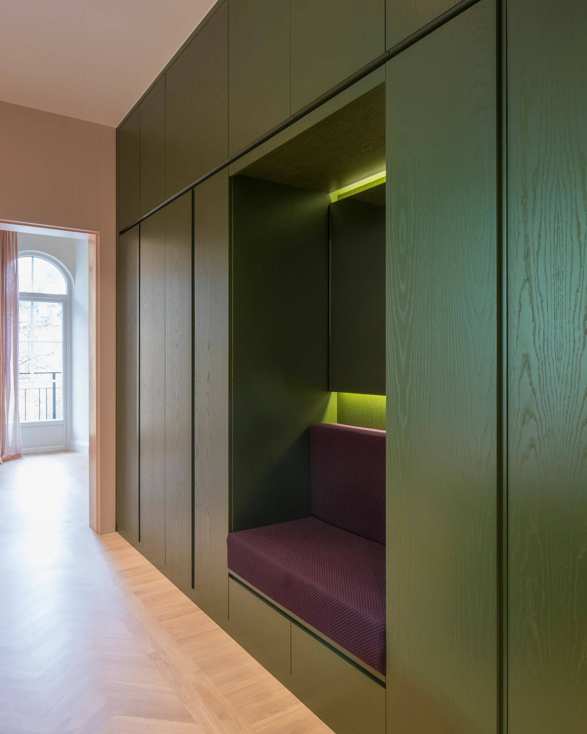 Floor-to-ceiling olive green cabinetry lines a hallway with a purple upholstered bench nook and light wood herringbone flooring.