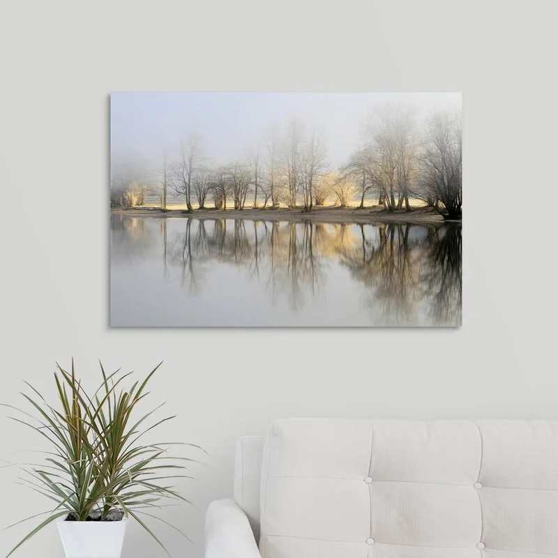 A cream tufted sofa sits beneath a large canvas print of misty trees reflected in water on a plain white wall next to a potted plant.