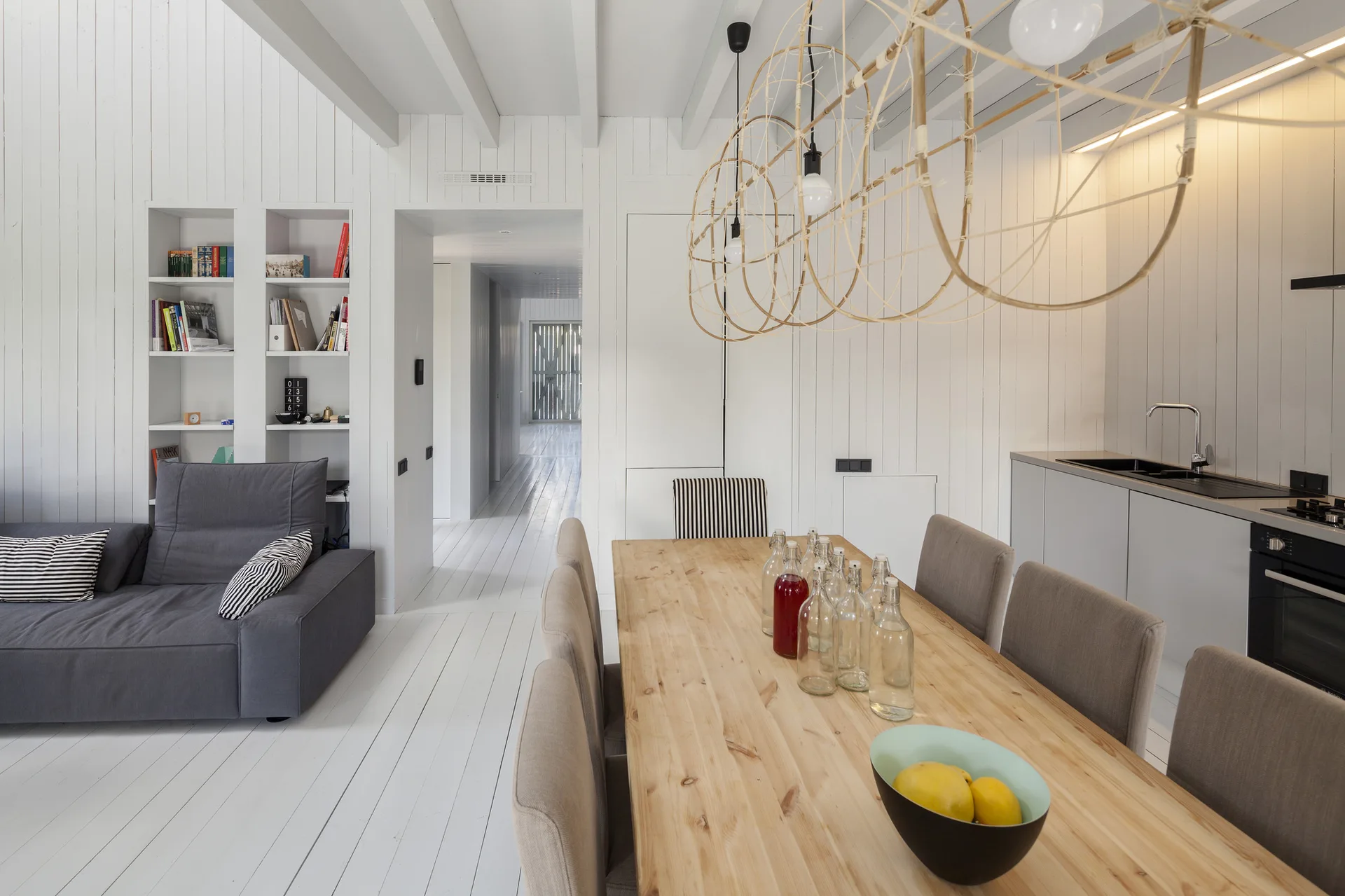 An open-plan room features white vertical wood paneling, a light wood dining table with grey chairs, and a grey sofa in the adjacent living area.