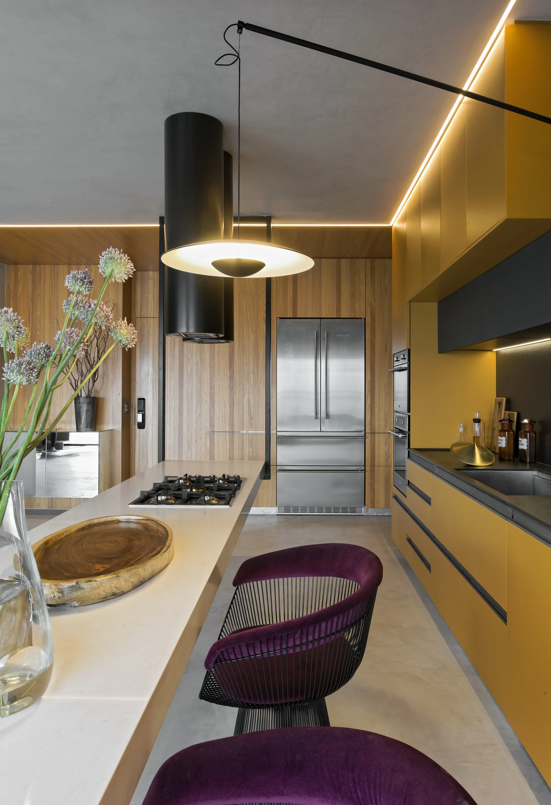 A modern kitchen with mustard yellow cabinets, wood paneled walls, a concrete floor, and purple velvet stools at the island.
