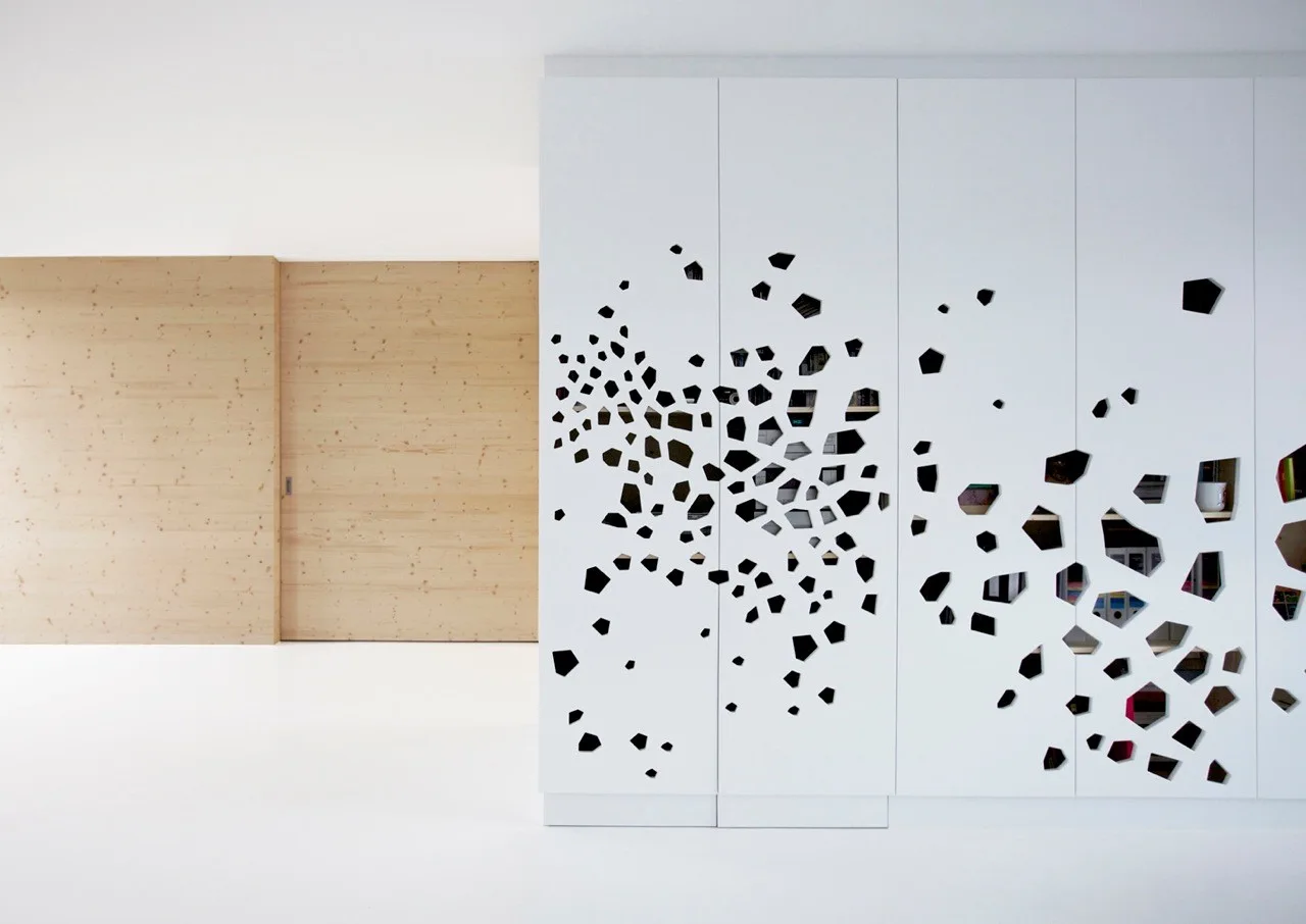 A minimalist white cabinet with irregular geometric cutouts stands next to a light wood panel sliding wall.