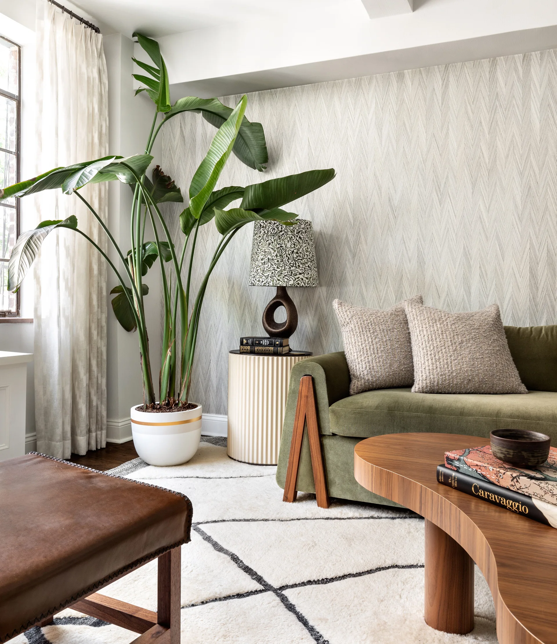 An olive green sofa sits next to a tall bird of paradise plant, a fluted side table, and a curved wood coffee table on a geometric area rug.
