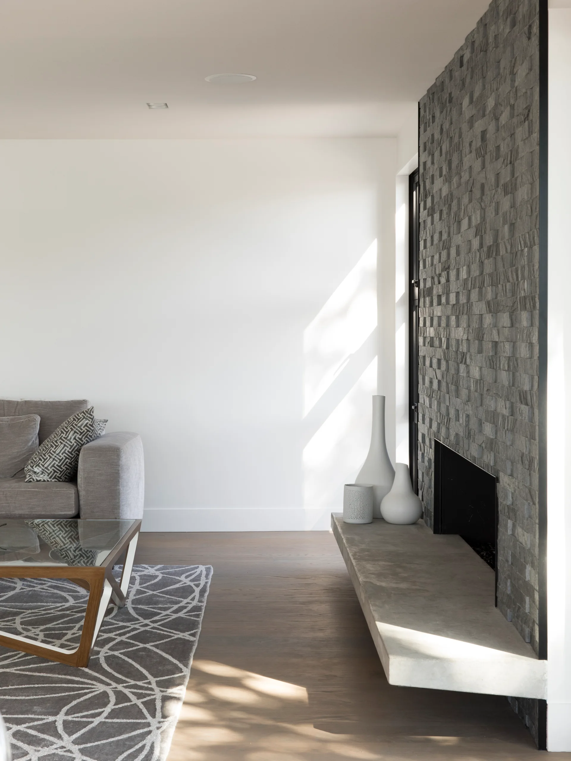 A modern living room features a dark stacked stone fireplace wall with a cantilevered concrete hearth, a grey sofa, and a patterned rug on wood flooring.