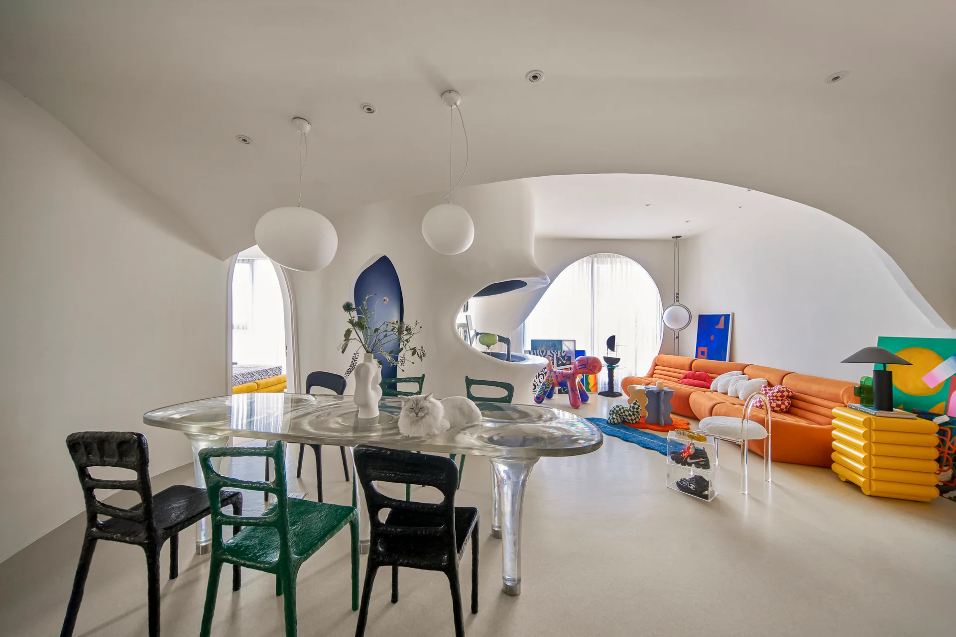 An open-plan room features organic white plaster walls, a translucent resin dining table, colorful sculptural chairs, and an orange channel-tufted sofa.