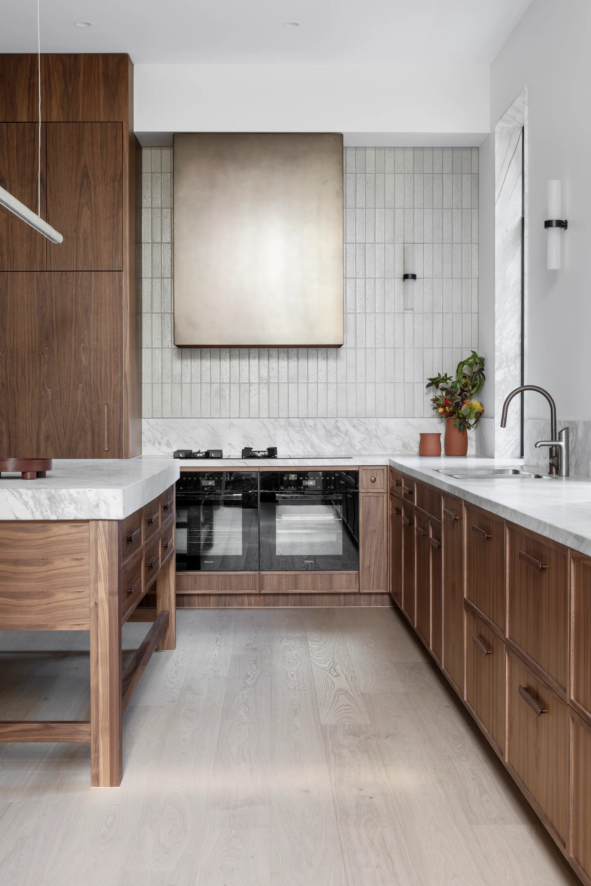 A modern kitchen featuring walnut wood cabinetry, a large metallic bronze range hood, white marble countertops, and a vertical stacked tile backsplash.