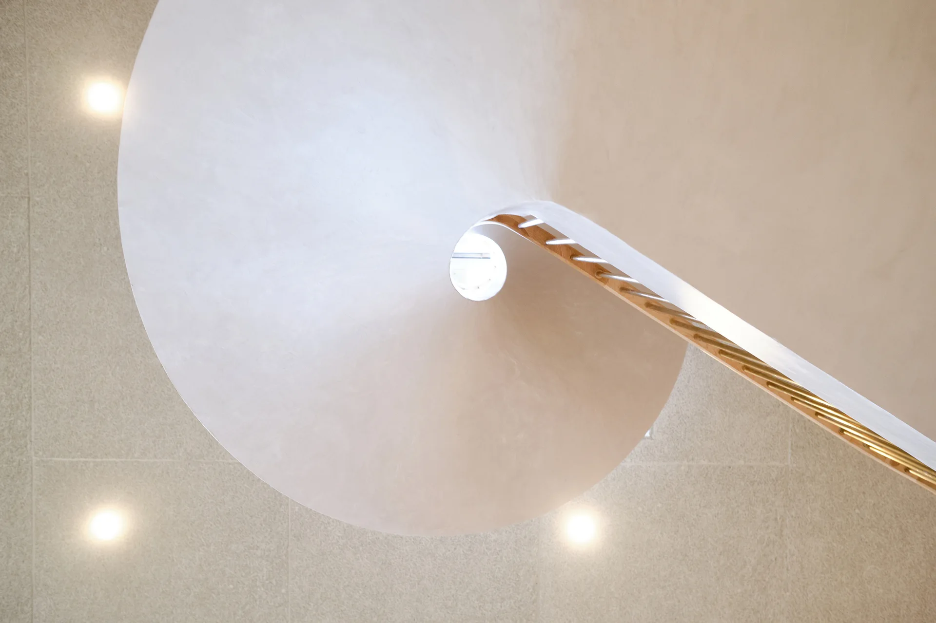 An upward-looking view captures the helical curve of a minimalist white spiral staircase with a wooden handrail against a beige textured ceiling.