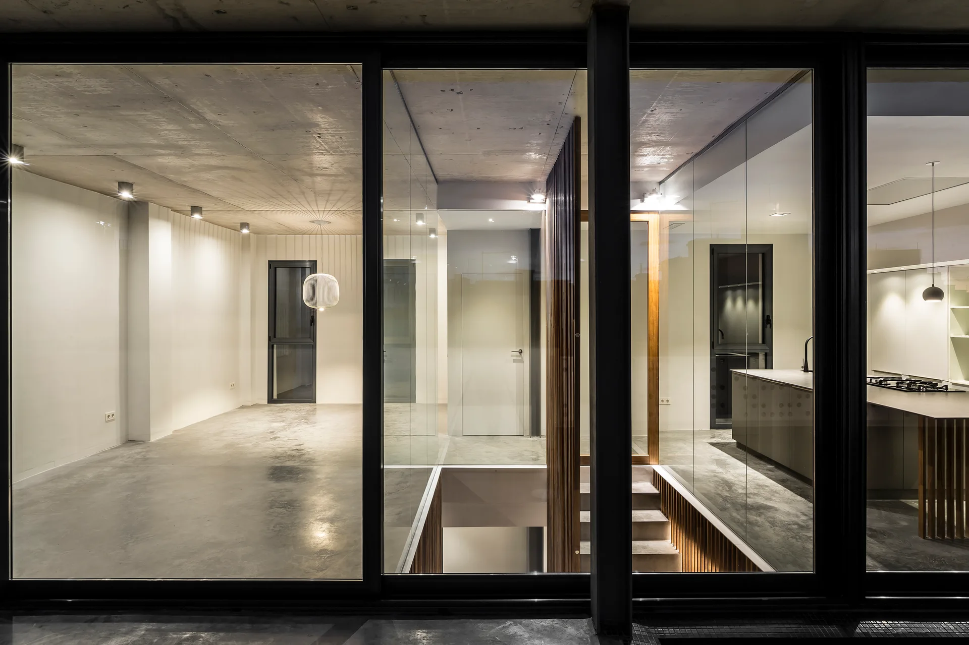Modern interior with glass partitions, concrete ceiling, staircase with wood slats, and a kitchen visible through windows.