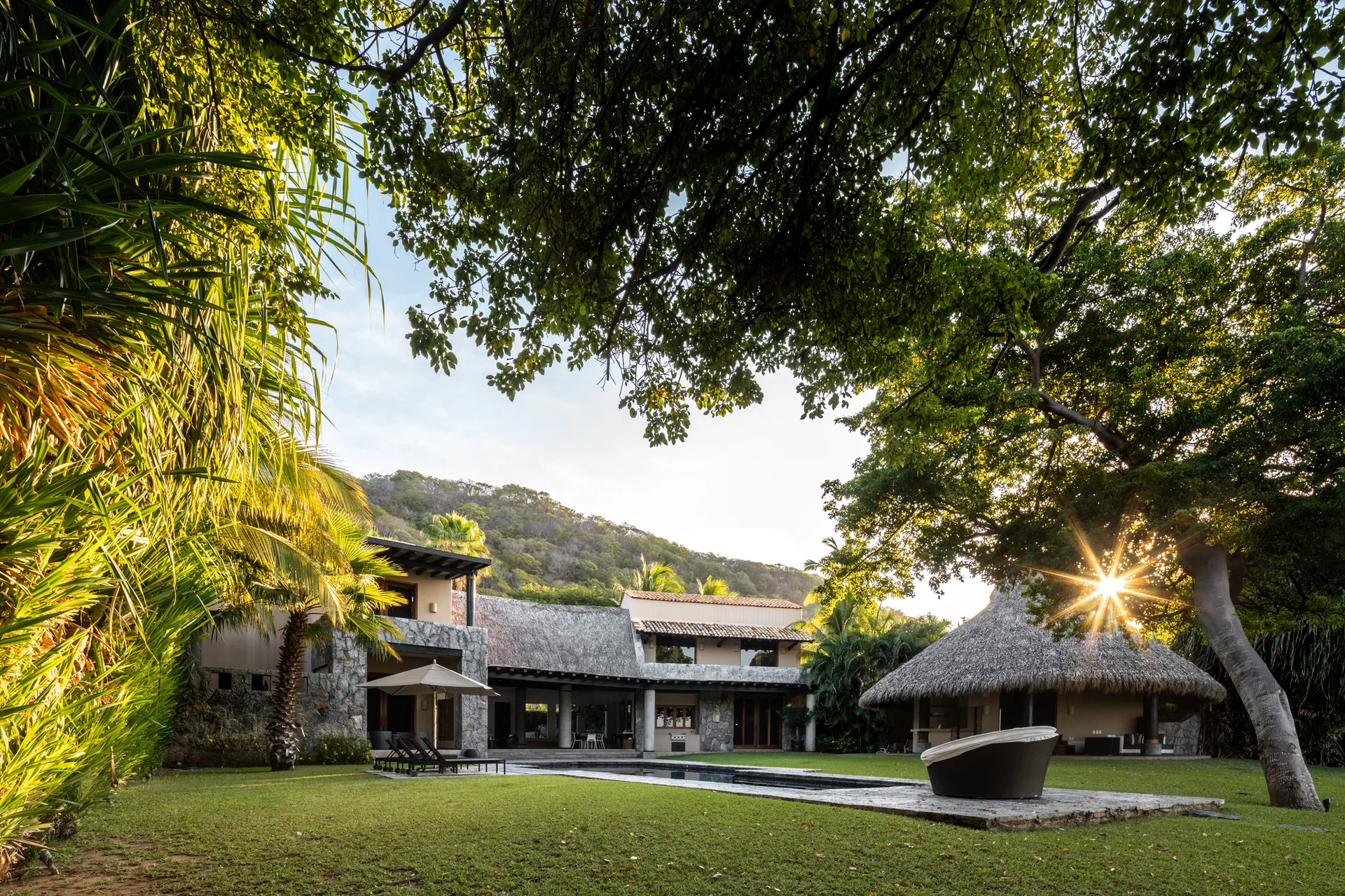A multi-level villa with a stone facade and thatched roof stands next to a swimming pool and a wide green lawn surrounded by tropical trees.