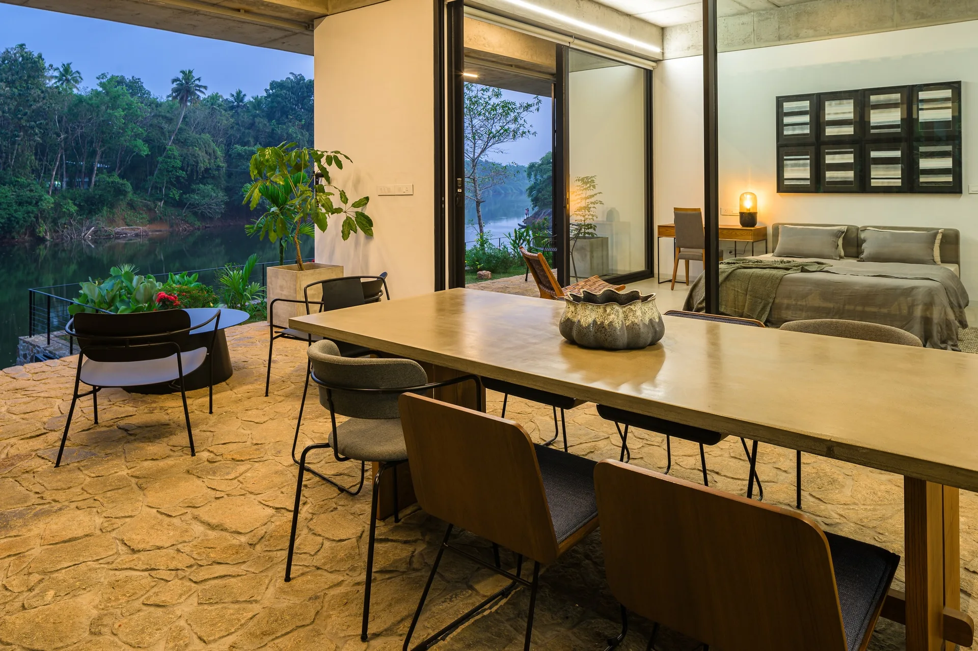 A covered outdoor patio features a large concrete dining table and mixed chairs set on flagstone flooring next to a glass-walled bedroom.