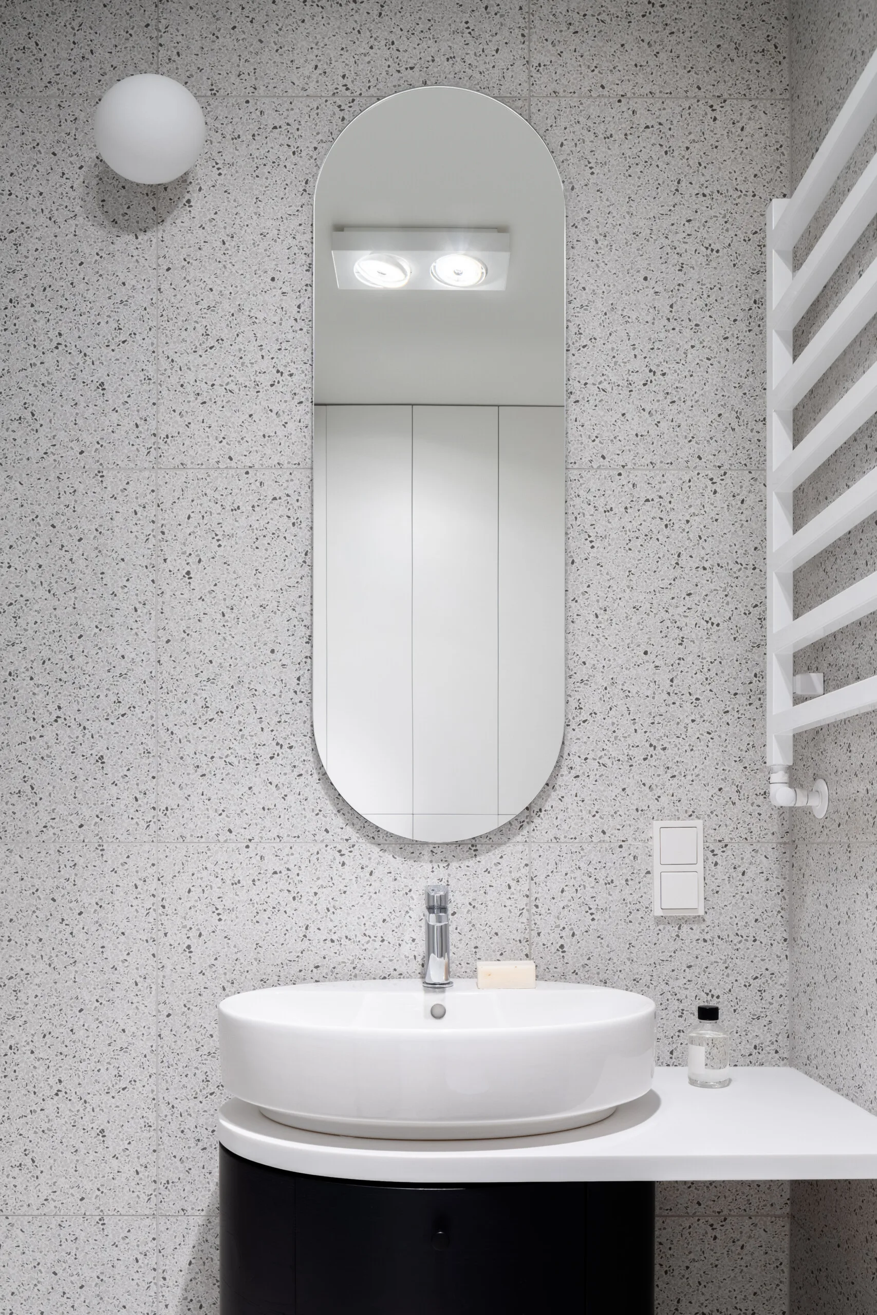 A white ceramic vessel sink rests on a black vanity against a grey terrazzo-tiled wall with a tall oval mirror.