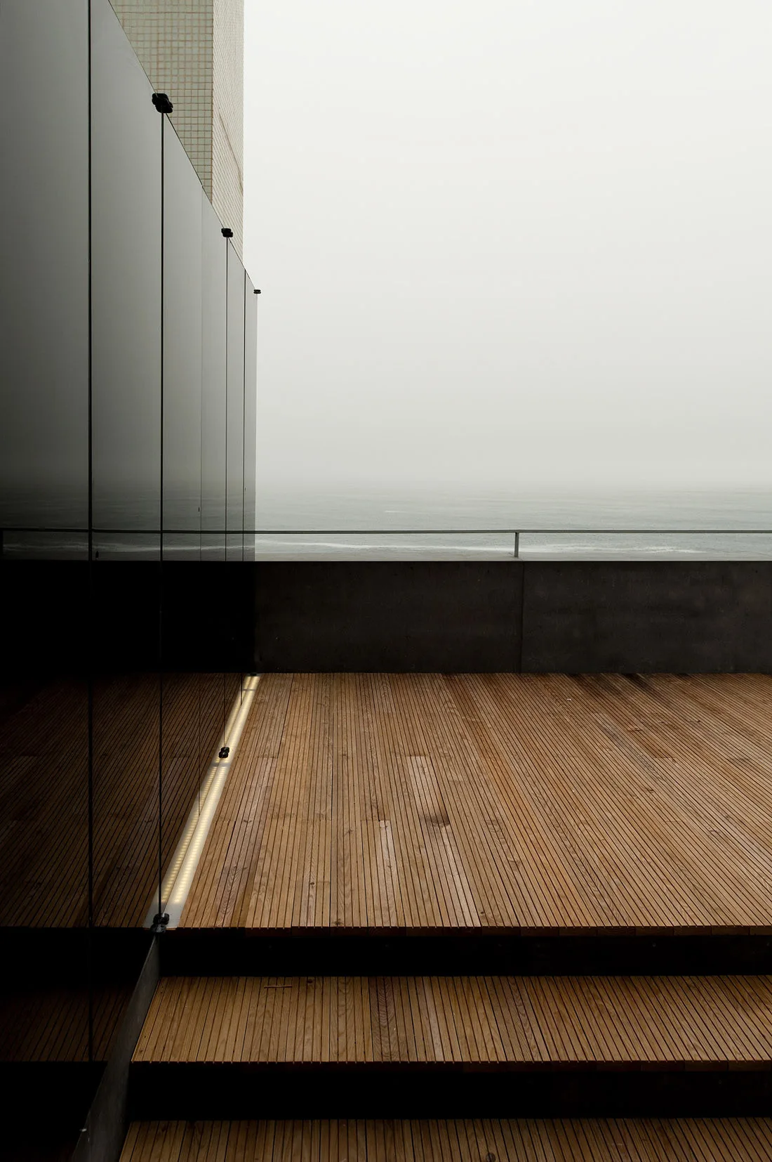 A modern outdoor wooden deck with a reflective glass wall, a horizontal metal railing, and a recessed linear floor light overlooking the ocean.