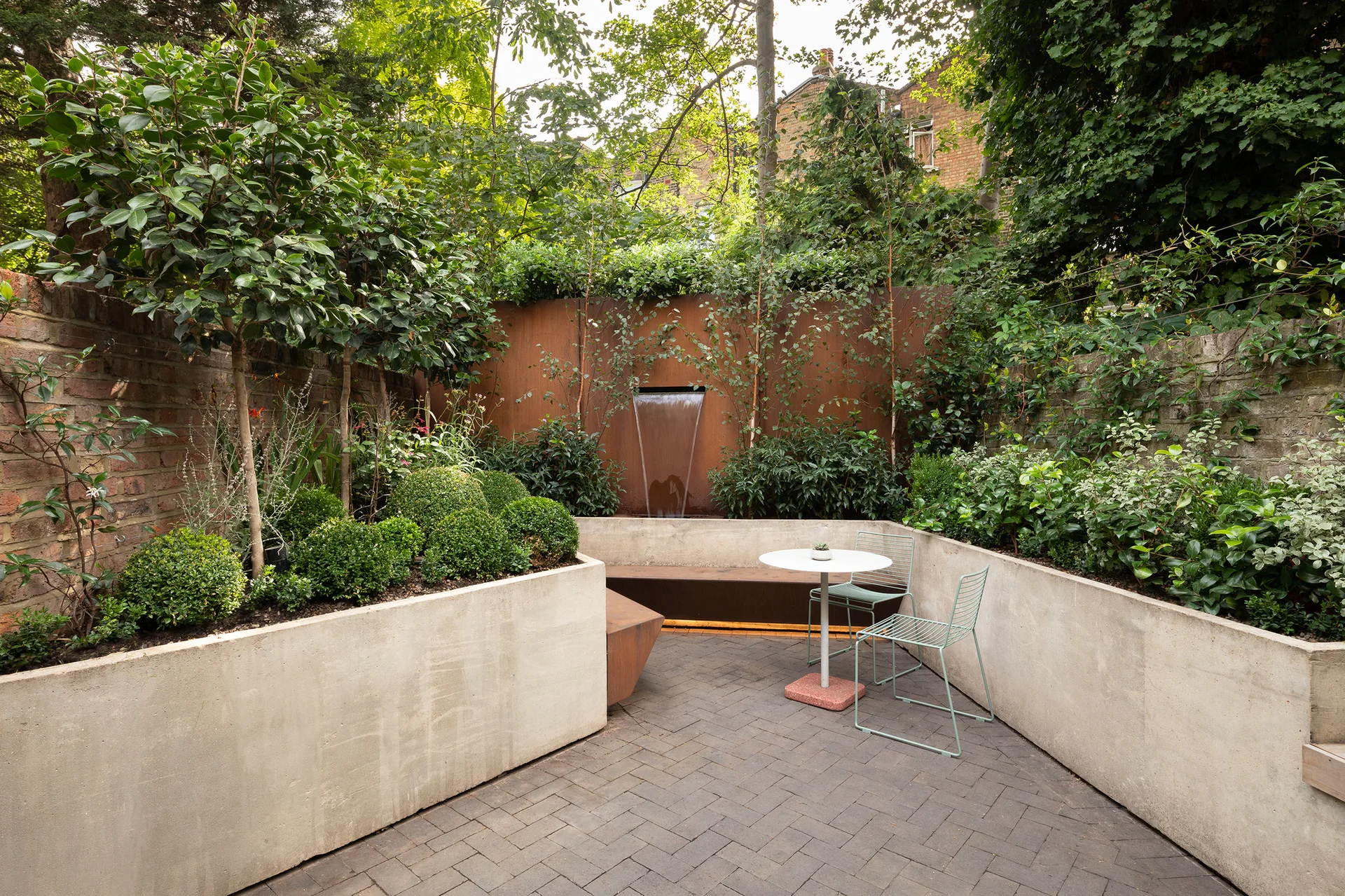 A modern courtyard garden features a rusted metal waterfall wall, poured concrete planters filled with greenery, and a bistro set on herringbone pavers.