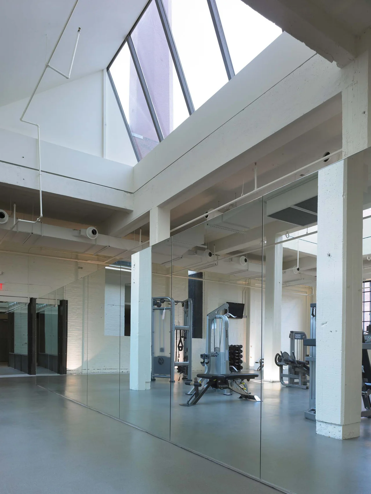 A spacious industrial gym featuring white structural columns, exercise equipment, full-height mirrored walls, and a large sloped skylight.