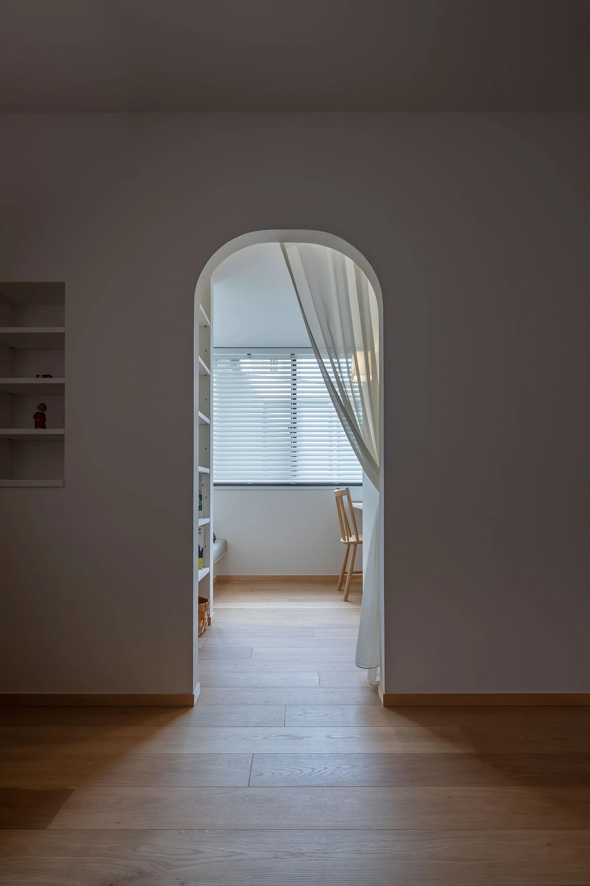A view through a white arched doorway into a sunlit minimalist room featuring wood floors, horizontal window blinds, and a sheer draped curtain.