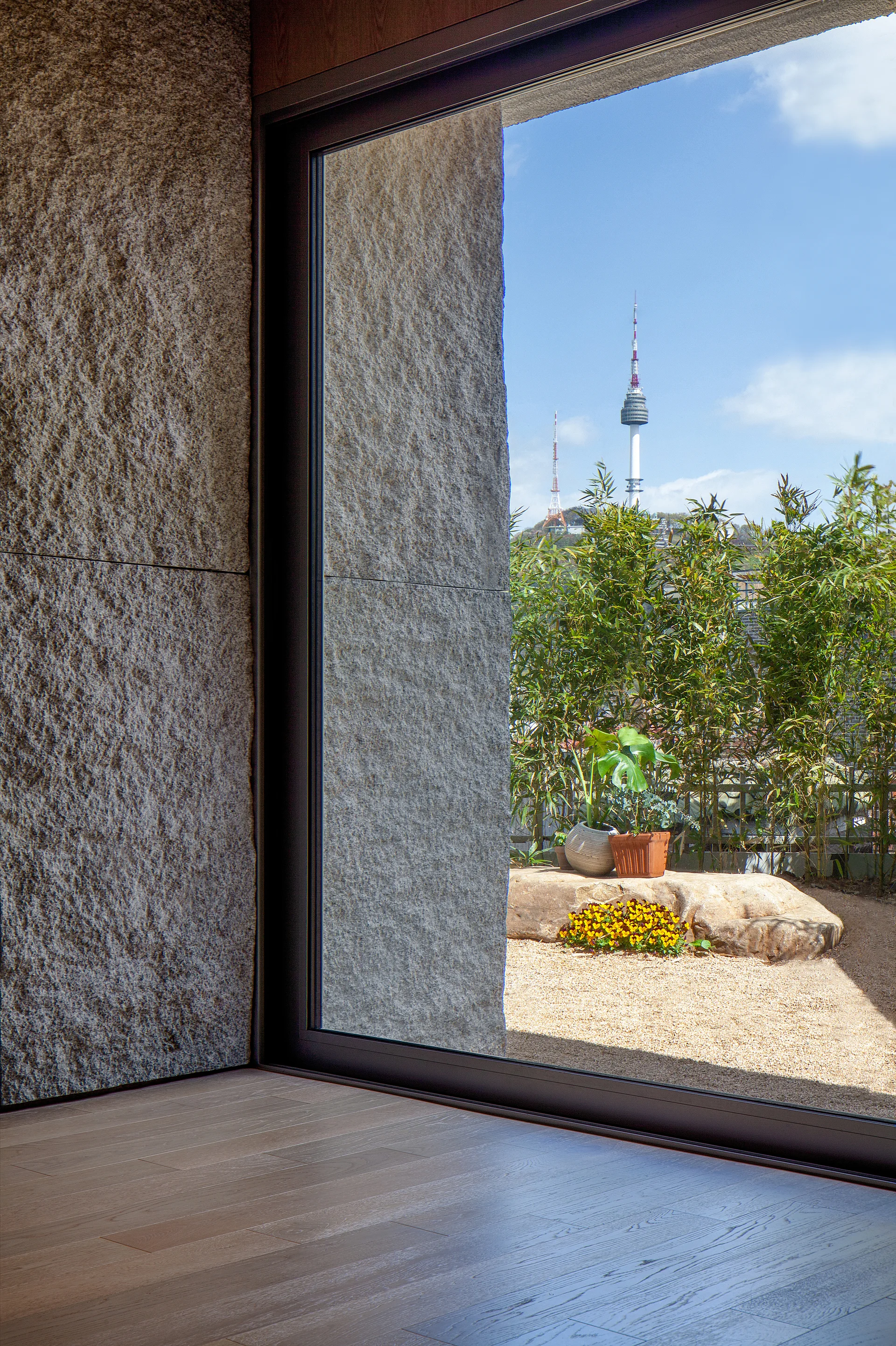 A textured grey stone wall and wood floor frame a large window looking out onto a gravel terrace with green bamboo and a distant landmark tower.