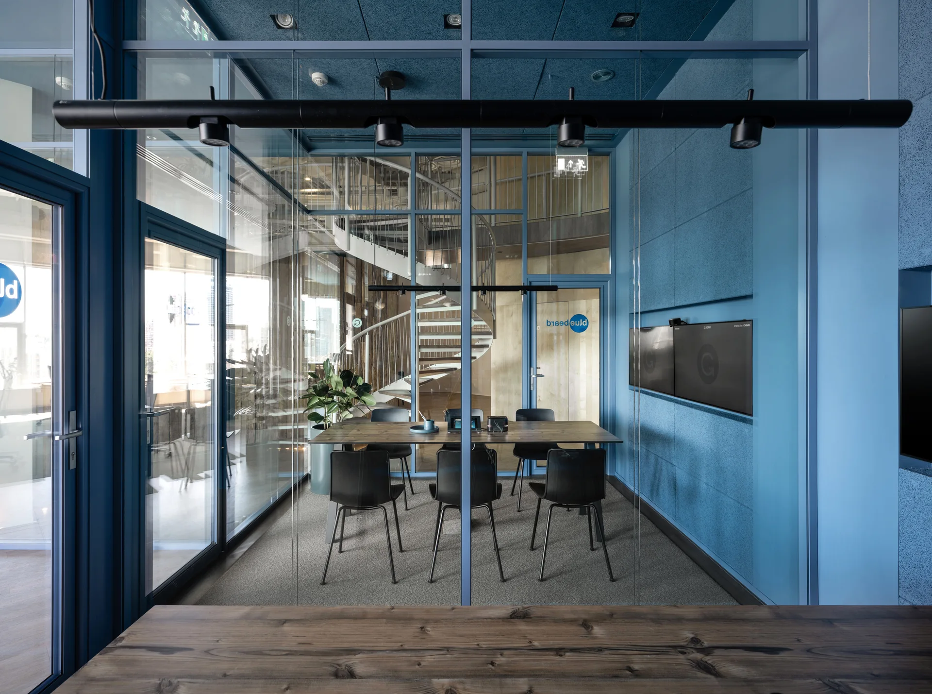 A modern blue-walled office meeting room features a central wood conference table, black chairs, and a glass partition showcasing a spiral staircase.