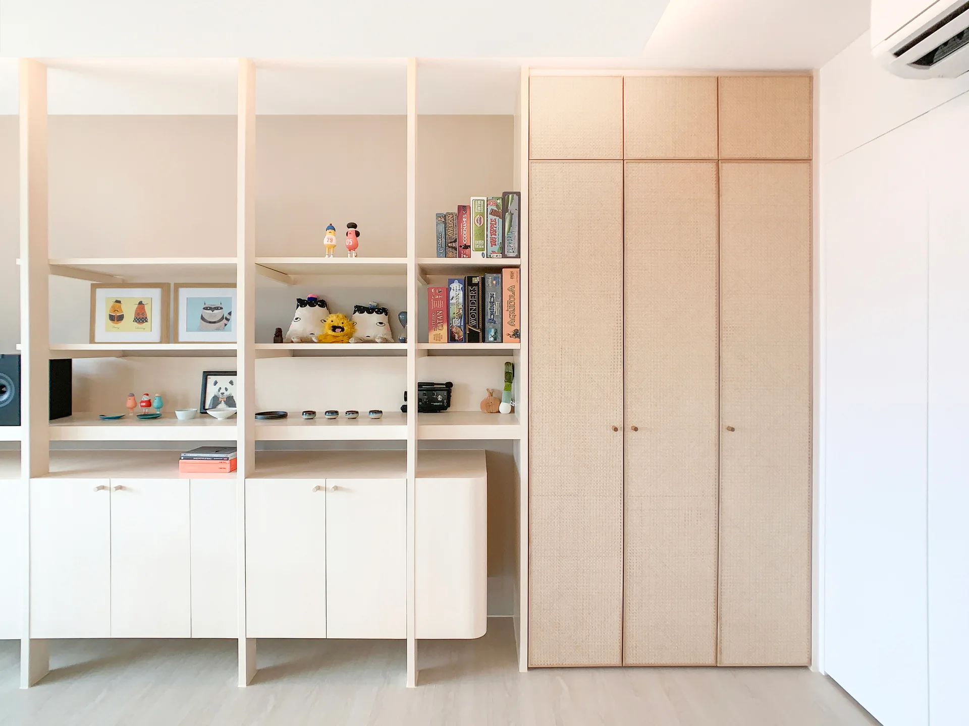 An off-white custom built-in shelving unit stands next to a tall closet with woven rattan doors on light wood flooring.