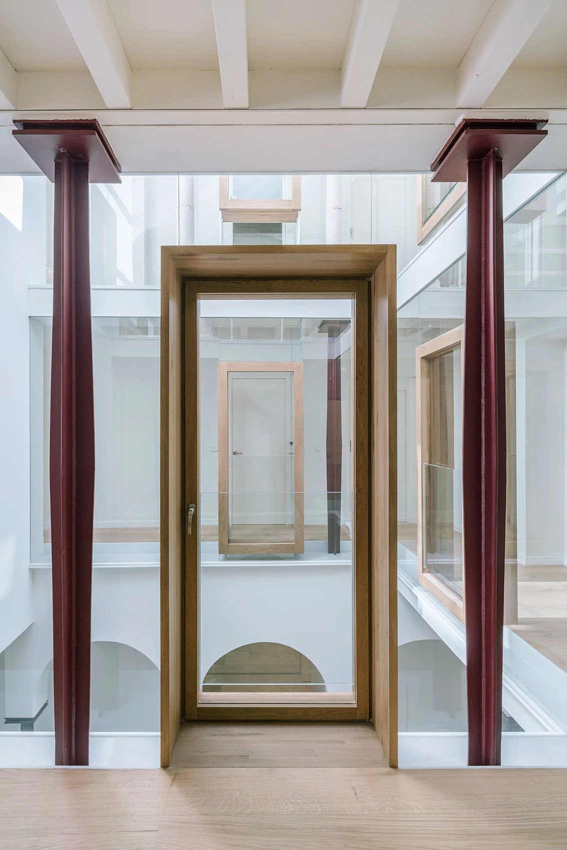 An interior hallway features structural red steel columns flanking a light wood door frame, with repeating wooden frames visible through glass partitions.