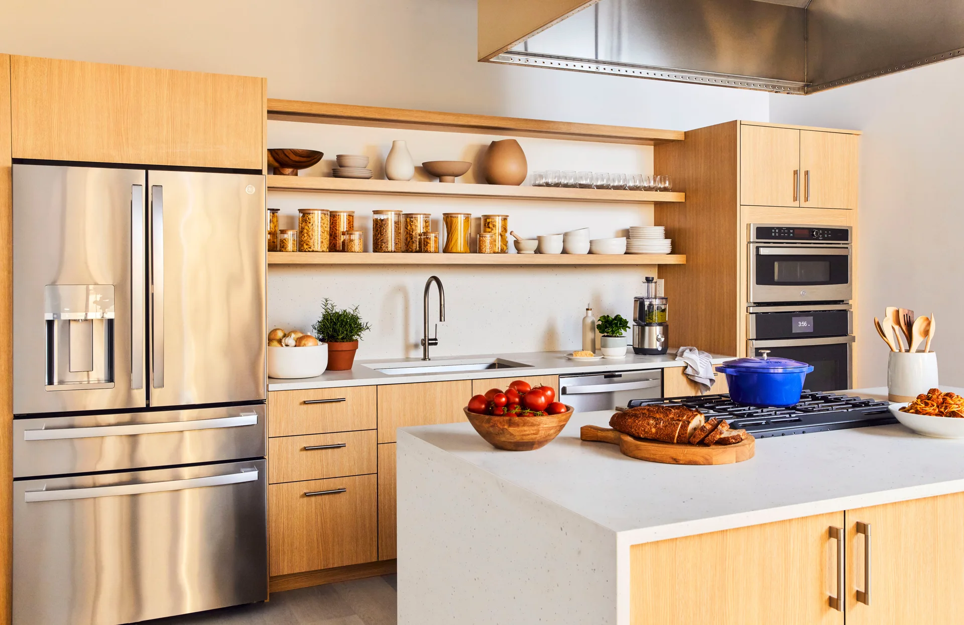 A modern kitchen with light wood flat-panel cabinets, open shelving holding jars and ceramics, stainless steel appliances, and a white stone island.