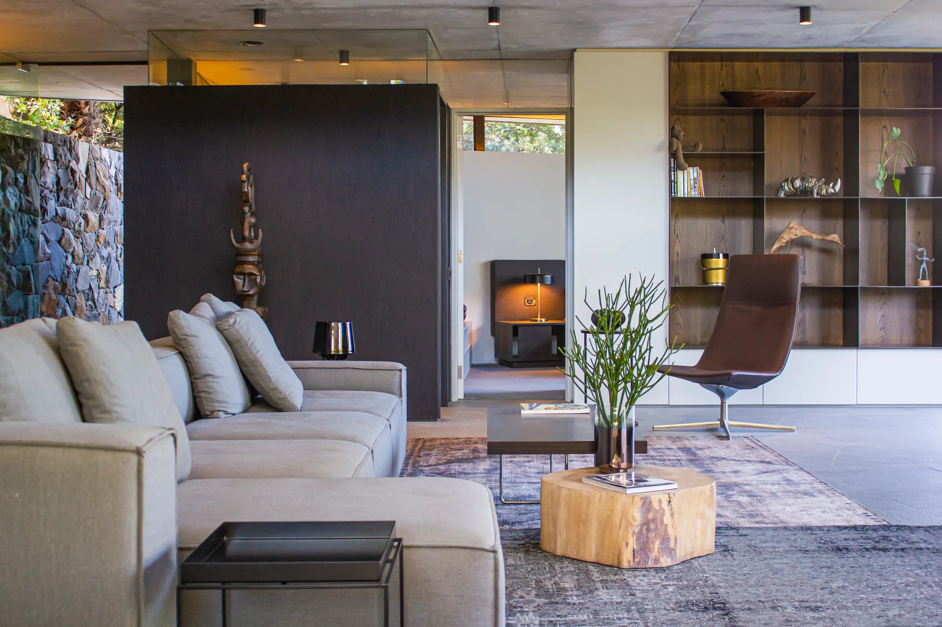 A modern living room features a grey sectional sofa, a brown leather swivel chair, an open wooden bookcase, and an exposed concrete ceiling.