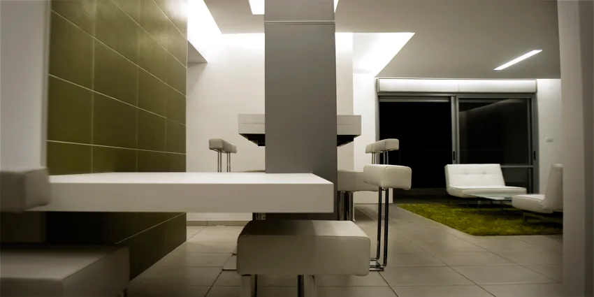 A minimalist dining area features a white table integrated into a grey central column next to a wall clad in olive green square tiles.