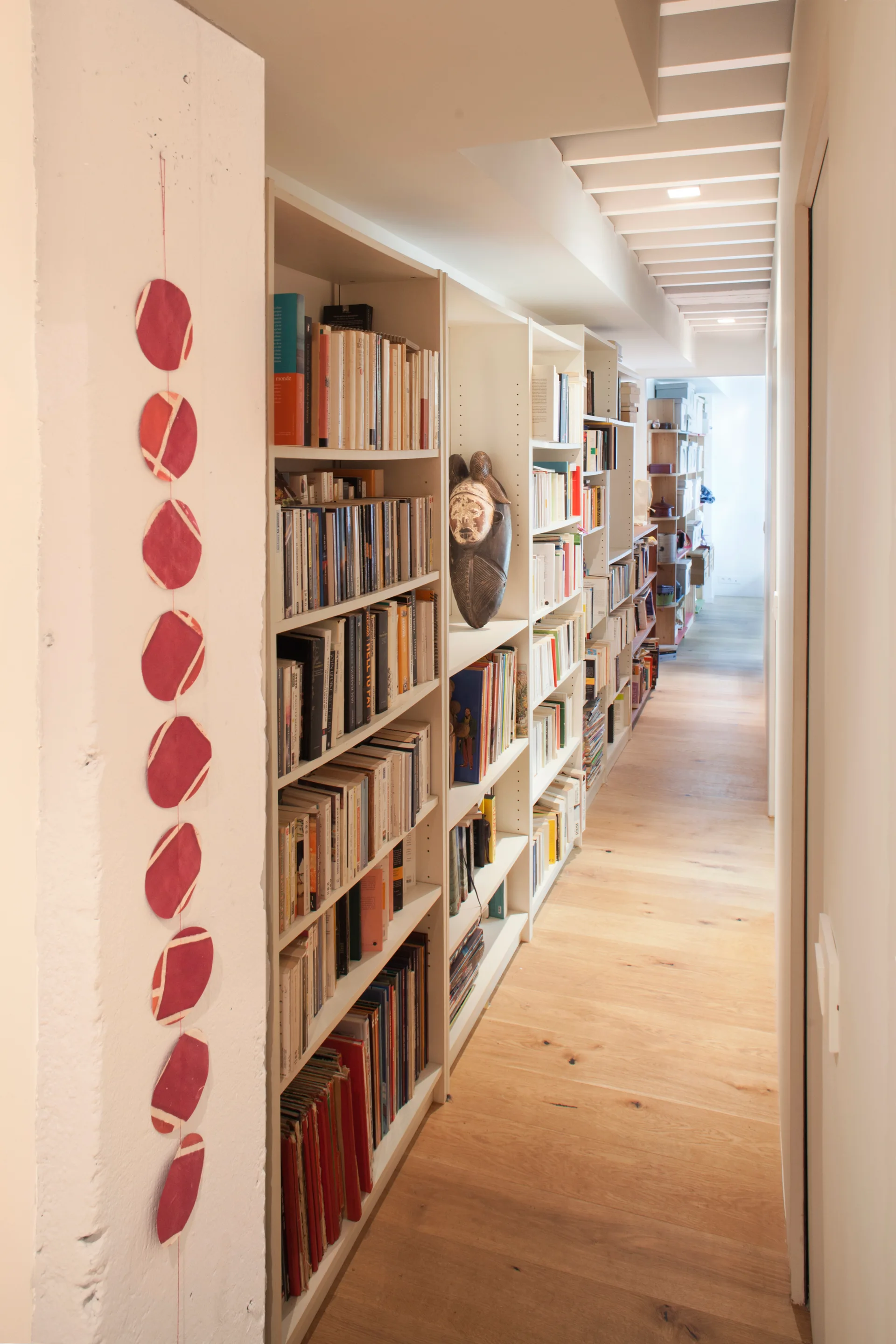 A narrow hallway featuring a long built-in white bookshelf filled with books next to light wood floors and an exposed beam ceiling.