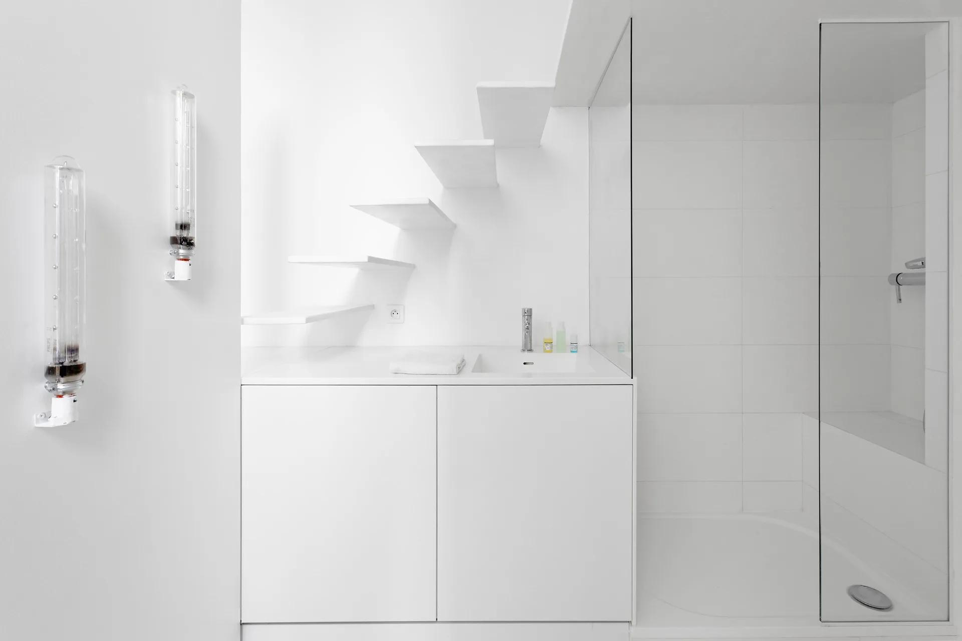 A minimalist white bathroom featuring a built-in vanity with a chrome faucet, vertical tube lights on the wall, and floating stair treads above.