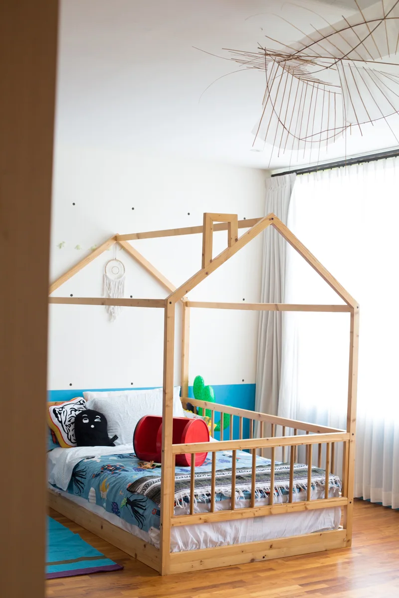 A children's bedroom features a natural wood house-shaped bed frame, blue and white painted walls, and a large sheer curtained window.