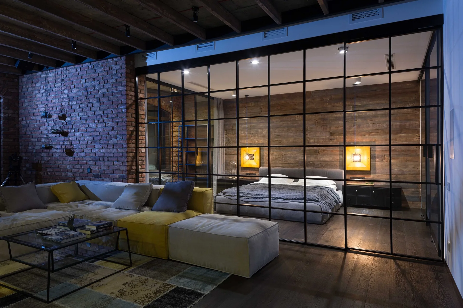 An industrial loft featuring a living room with a sectional sofa and a bedroom visible behind a black metal grid glass partition wall.