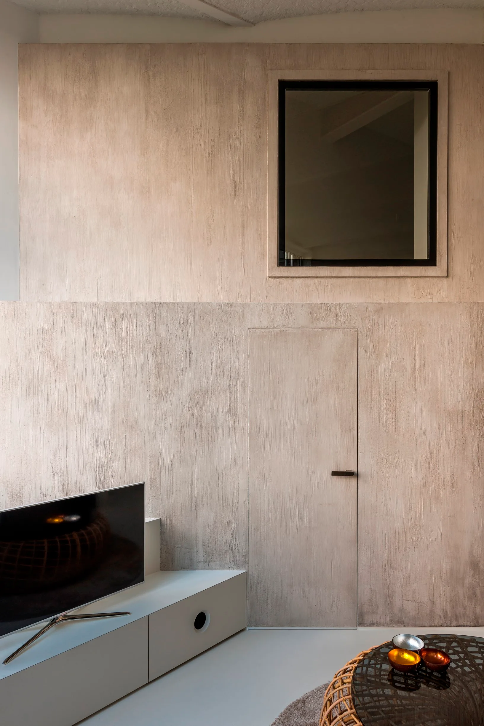 A minimalist living room features a textured greige plaster wall with an integrated flush door, a small high window, and a low white media console.