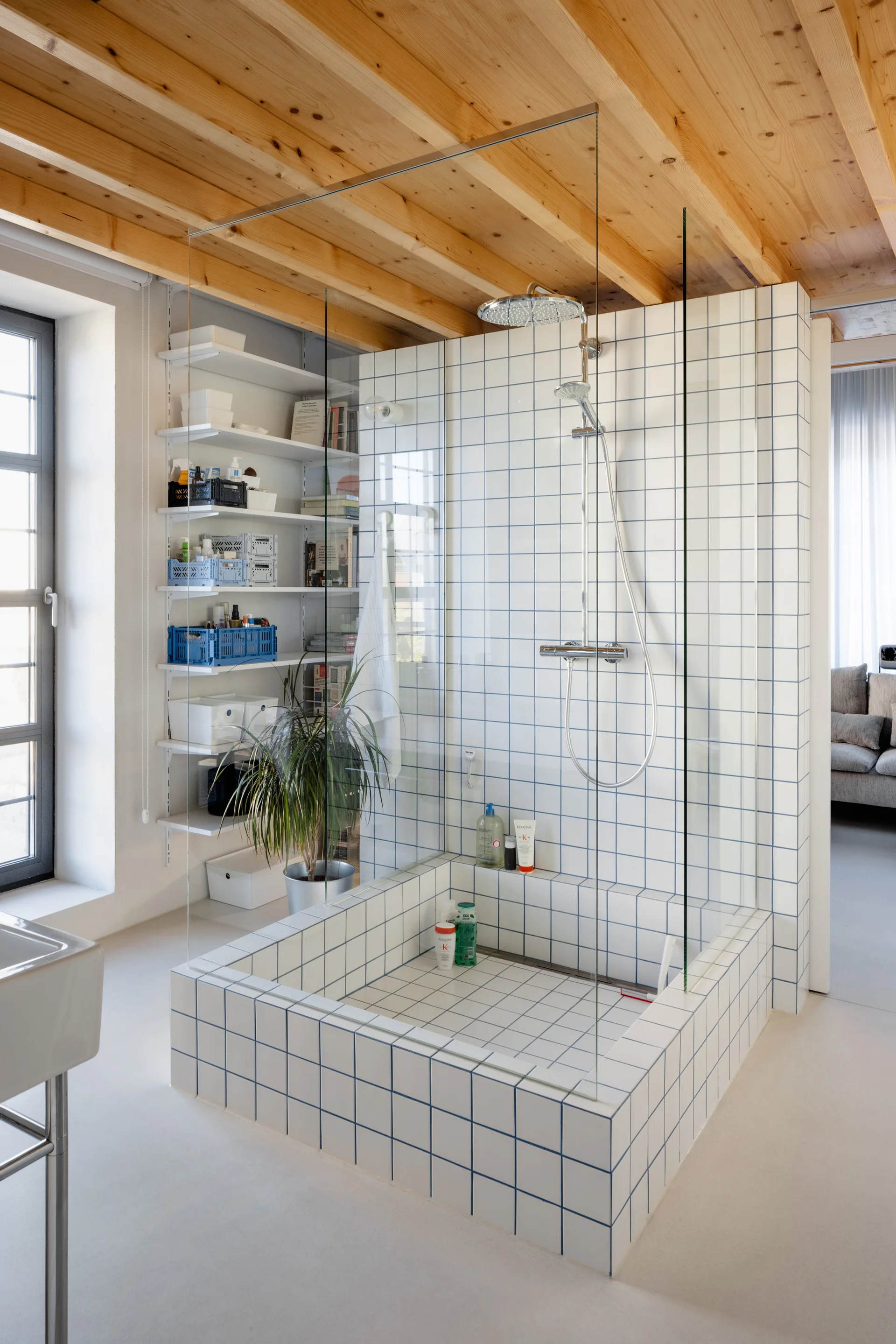 A modern walk-in shower enclosed in frameless glass features square white tiles with blue grout beneath a natural wood plank ceiling.