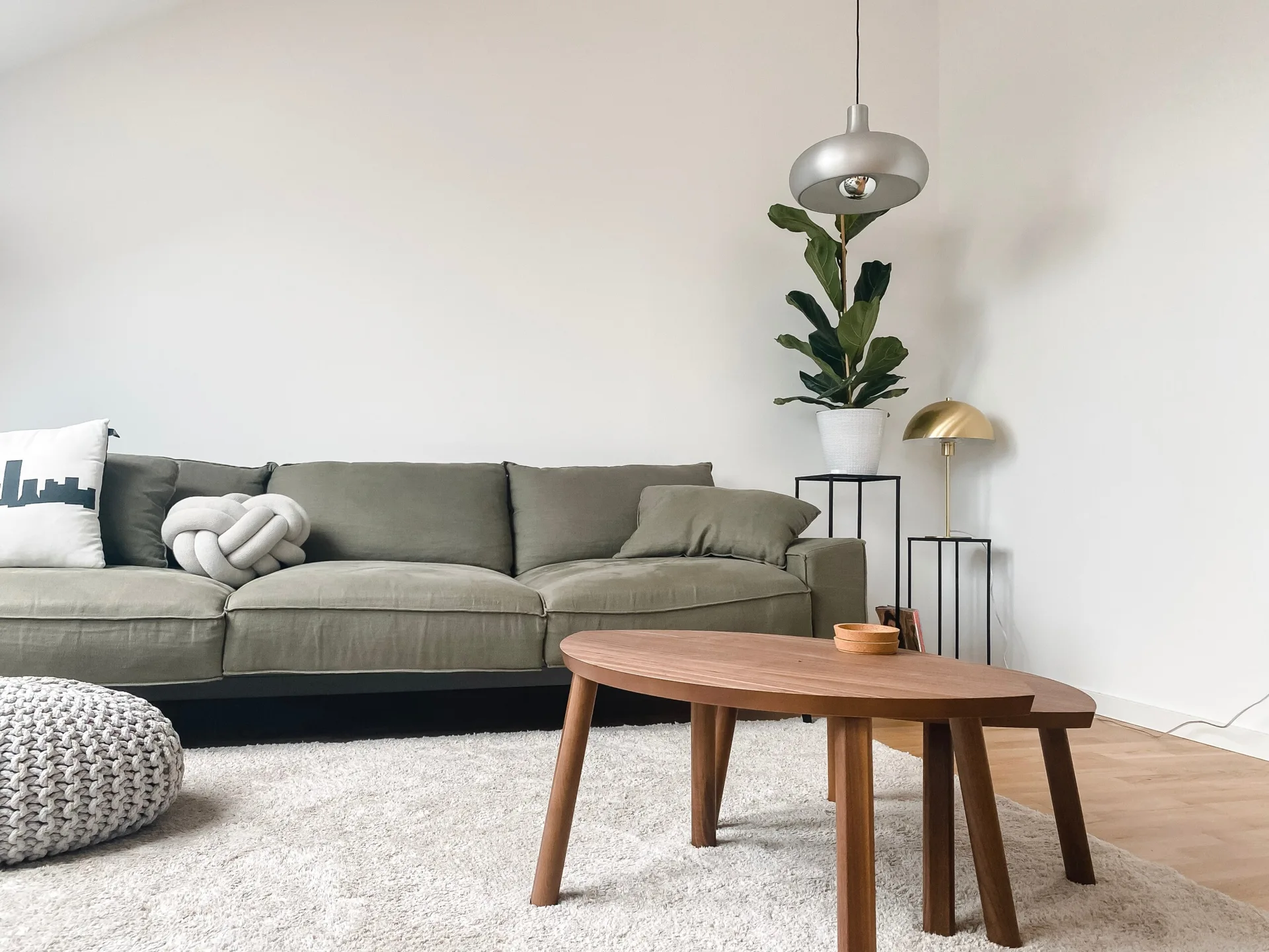 An olive green sofa sits on a textured white rug next to nested wooden coffee tables, accompanied by a potted plant and a metal pendant lamp.