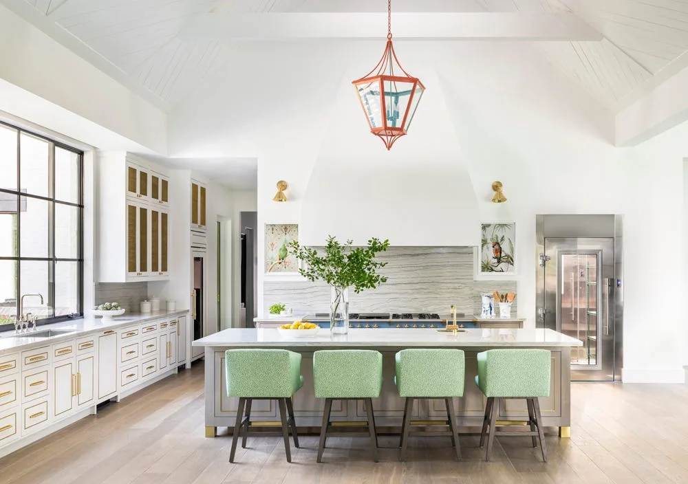 A bright kitchen features a large central island with four green counter stools, a white custom range hood, and an orange lantern pendant light.