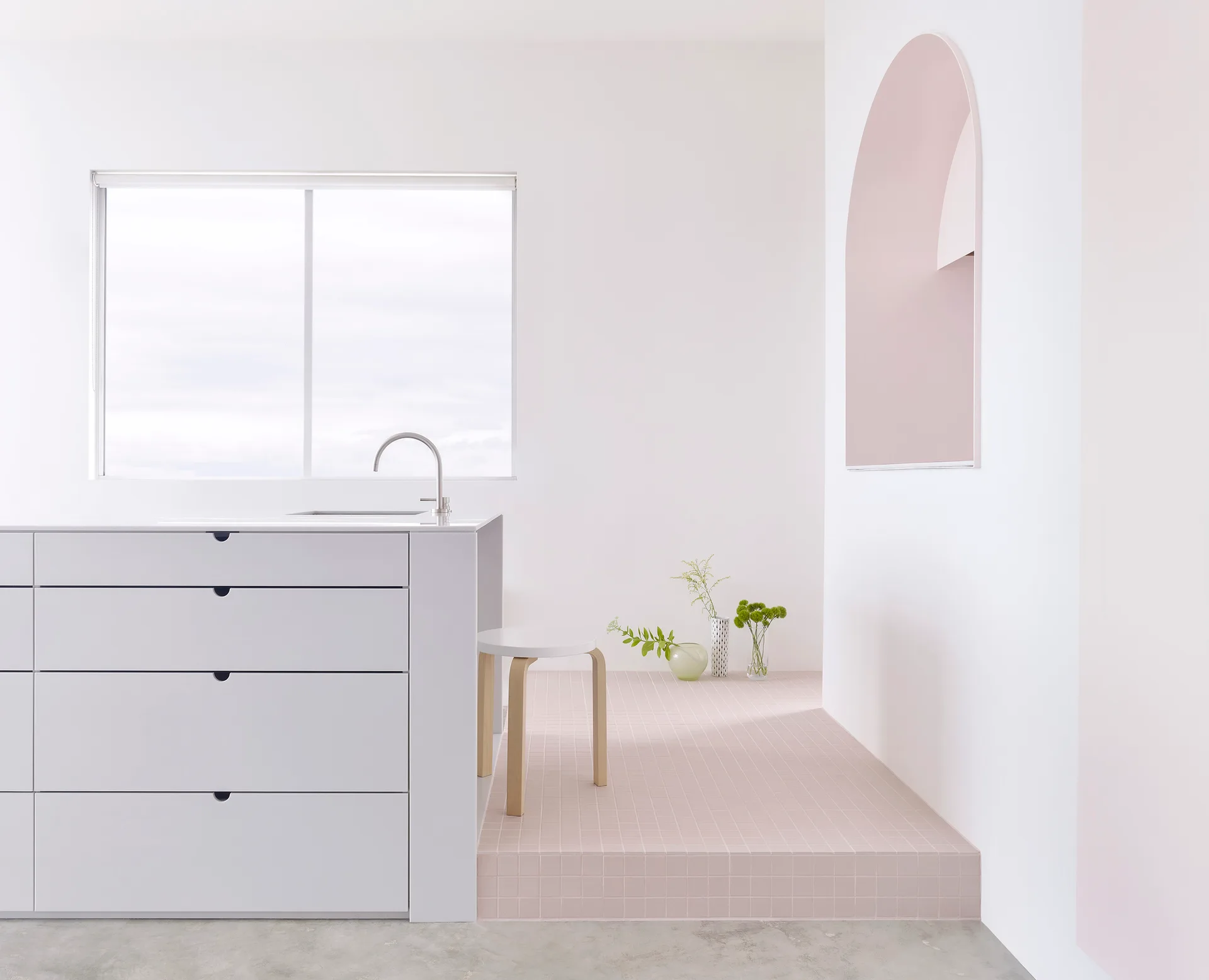 A minimalist white kitchen features a large drawer island next to a raised pink tiled platform with an arched niche in the wall.