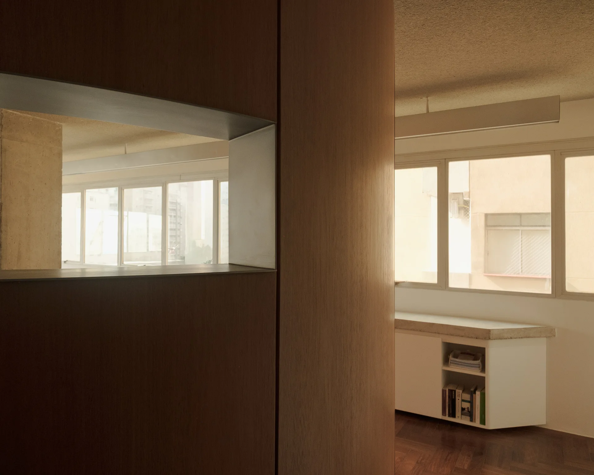 A wood-paneled partition wall featuring a rectangular cutout stands near a white low cabinet and large apartment windows.