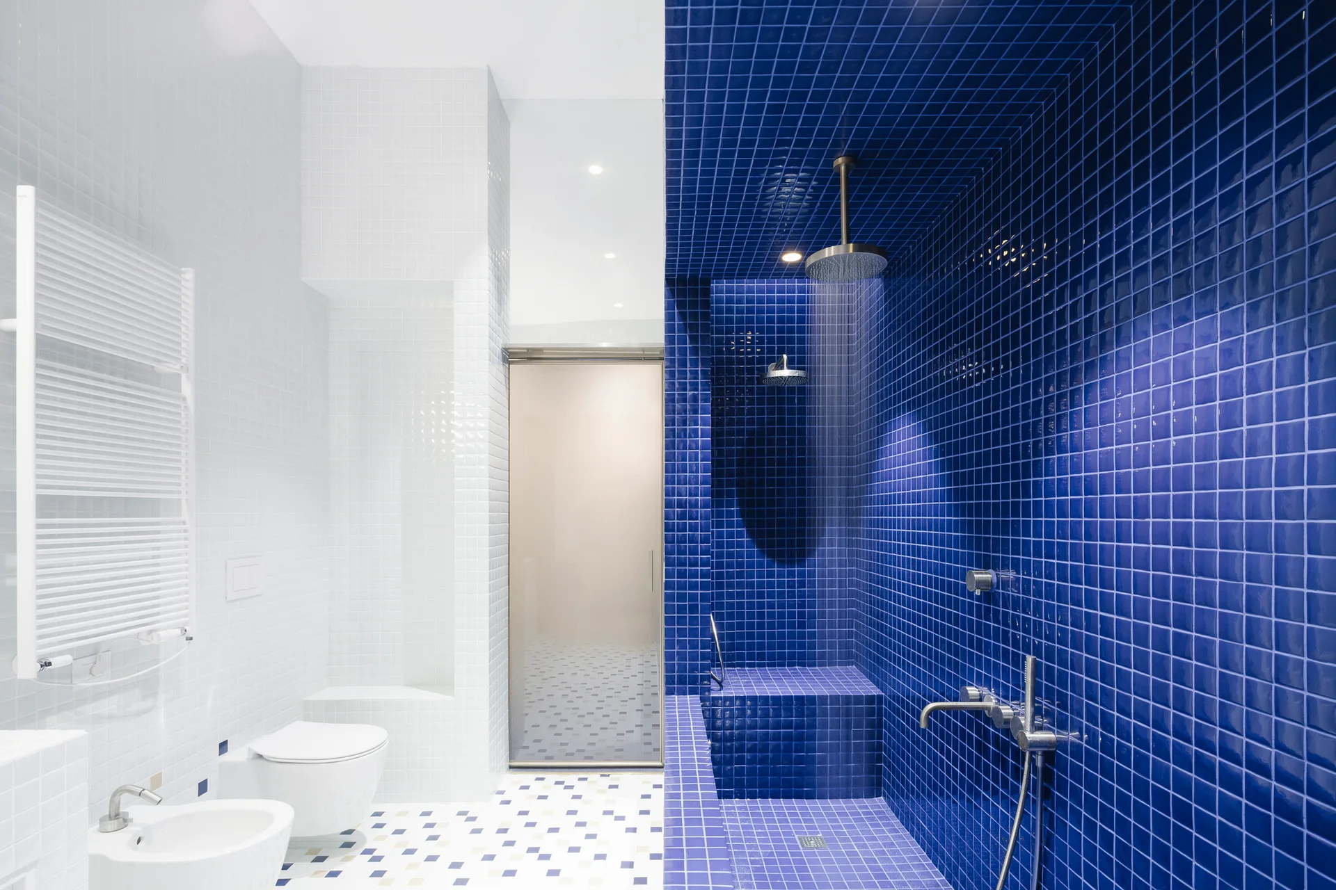 A modern bathroom with white square tiles on one side and a cobalt blue tiled walk-in shower with a built-in bench on the other.