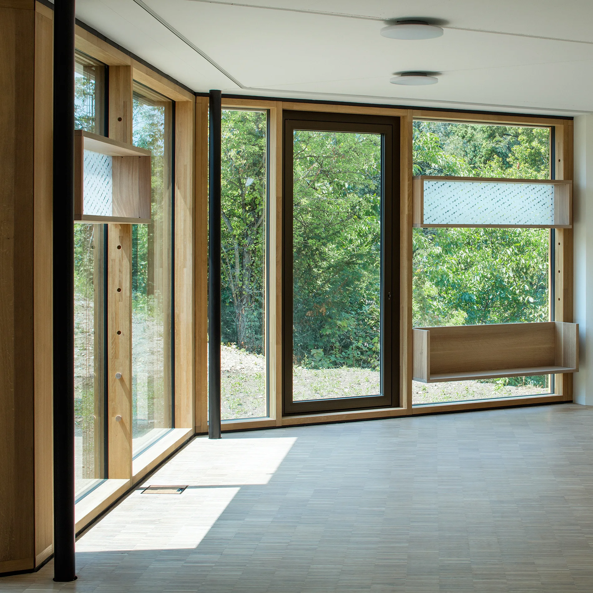 Large floor-to-ceiling windows with thick light wood frames feature built-in wooden box shelves and look out onto green trees.