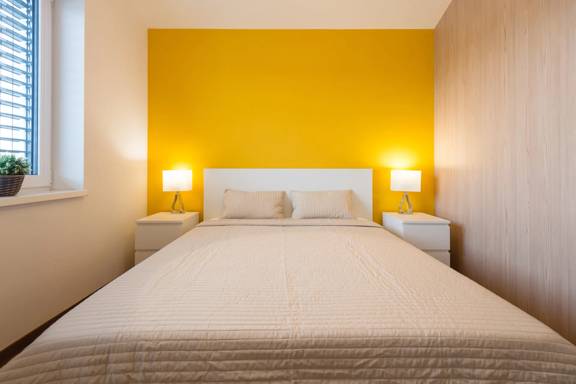 A minimalist bedroom with a bright yellow accent wall behind a white bed, flanked by two matching nightstands with glowing table lamps.