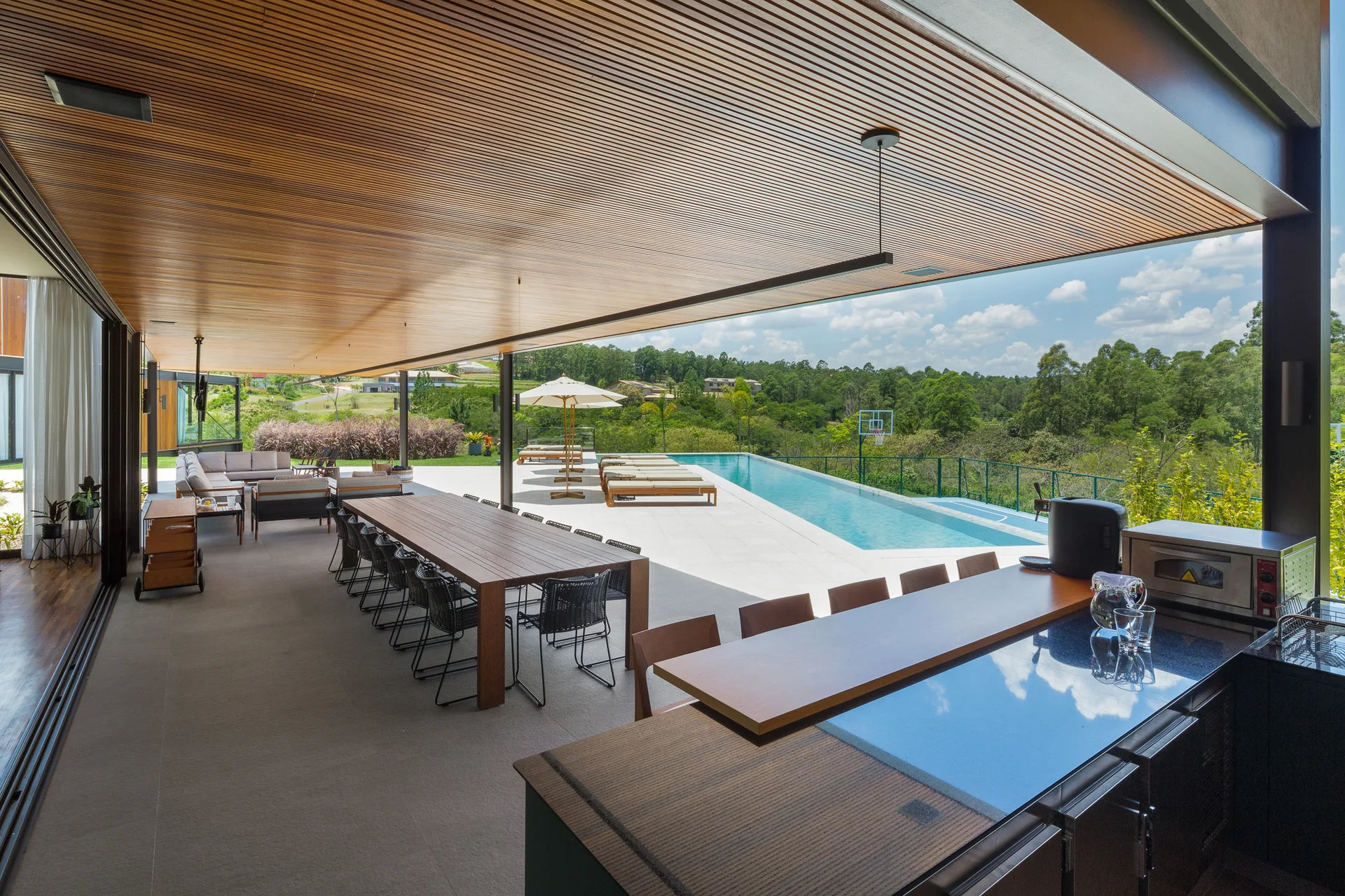 A spacious covered patio features a slatted wood ceiling, a long wooden dining table, an outdoor kitchen counter, and a nearby swimming pool.