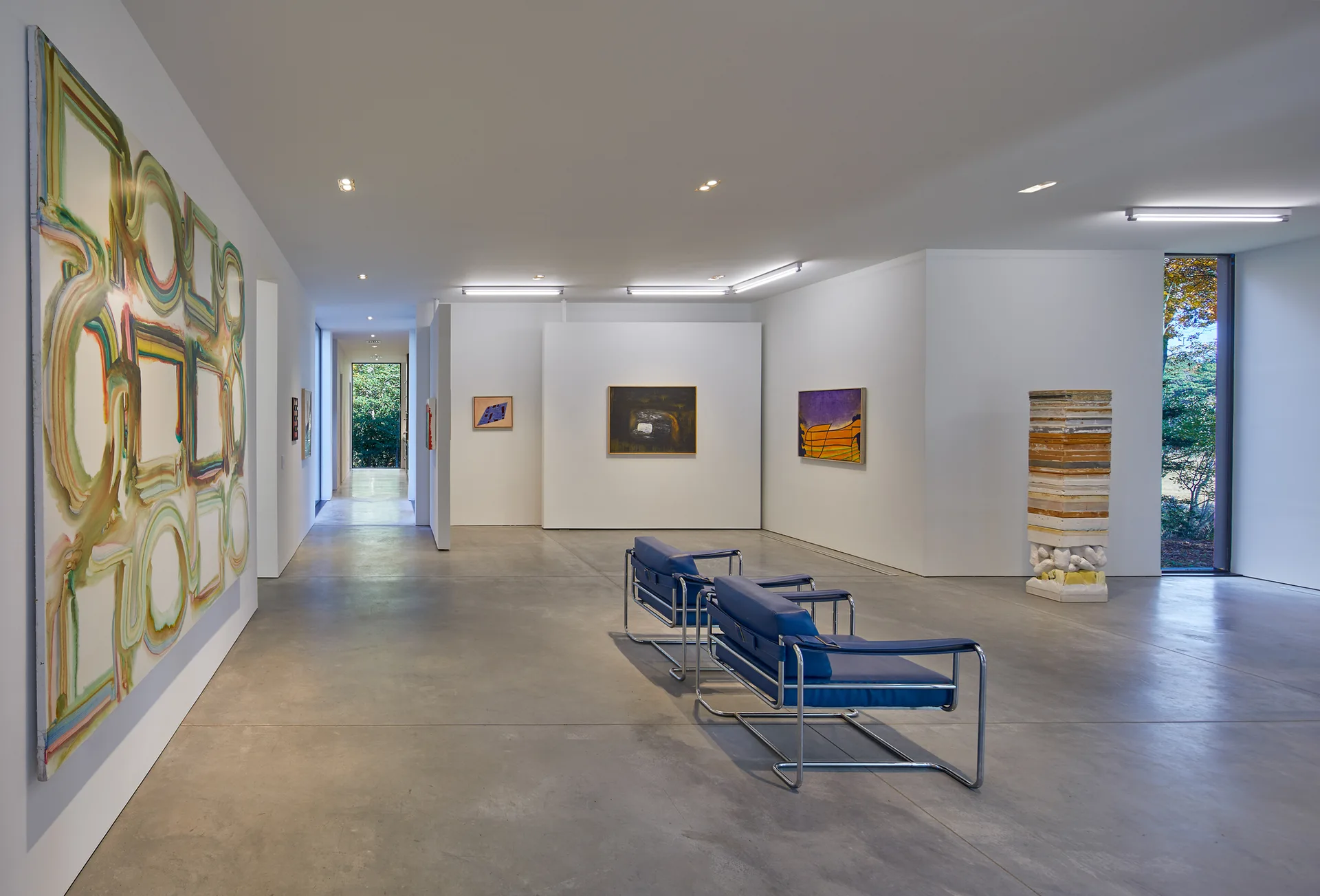 A minimalist art gallery features white walls with framed paintings, polished concrete floors, and a pair of blue tubular steel chairs.