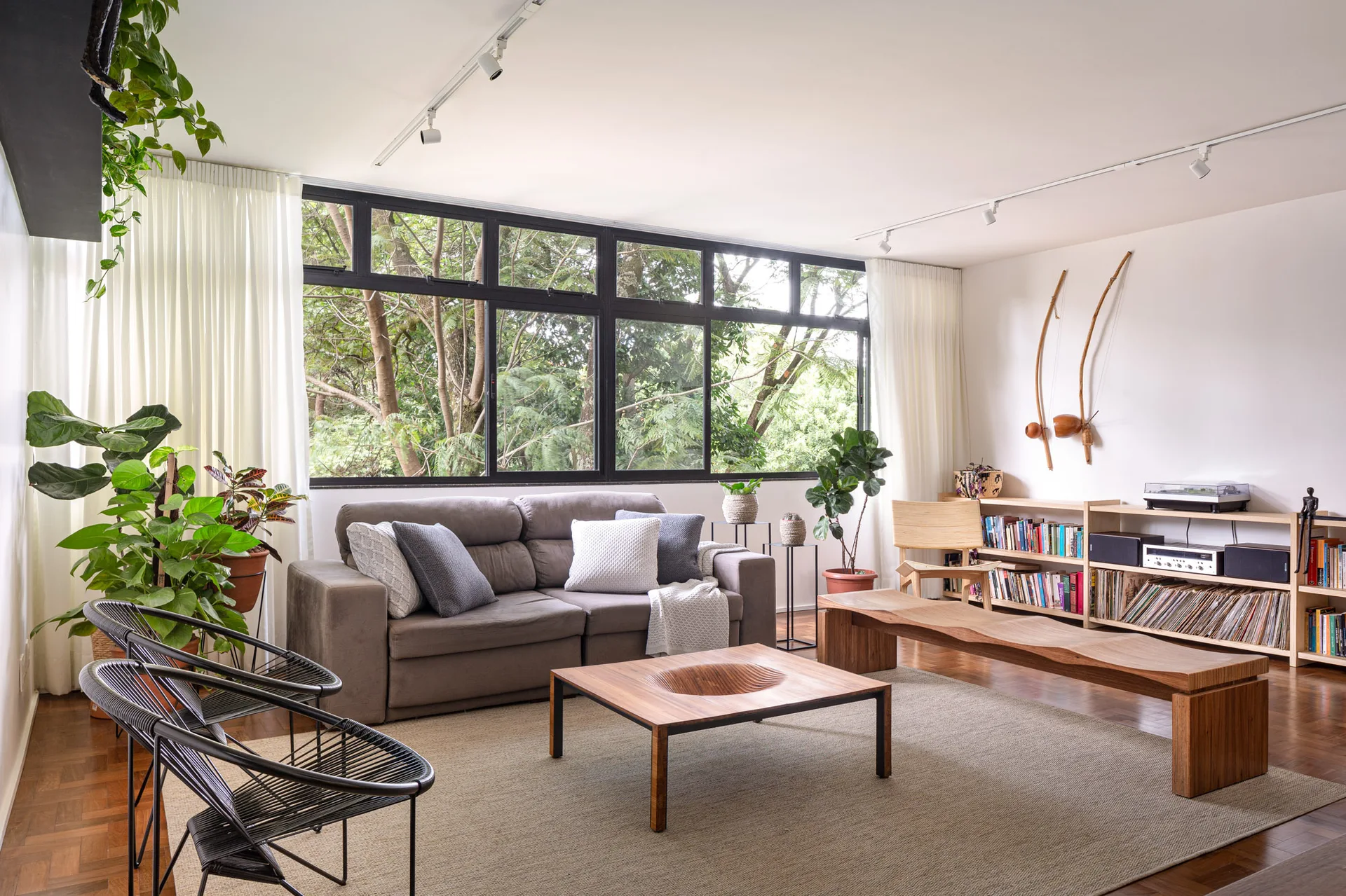 A mid-century living room features a grey sofa, a carved wooden coffee table, a long wooden bench, and a large window looking out at green trees.