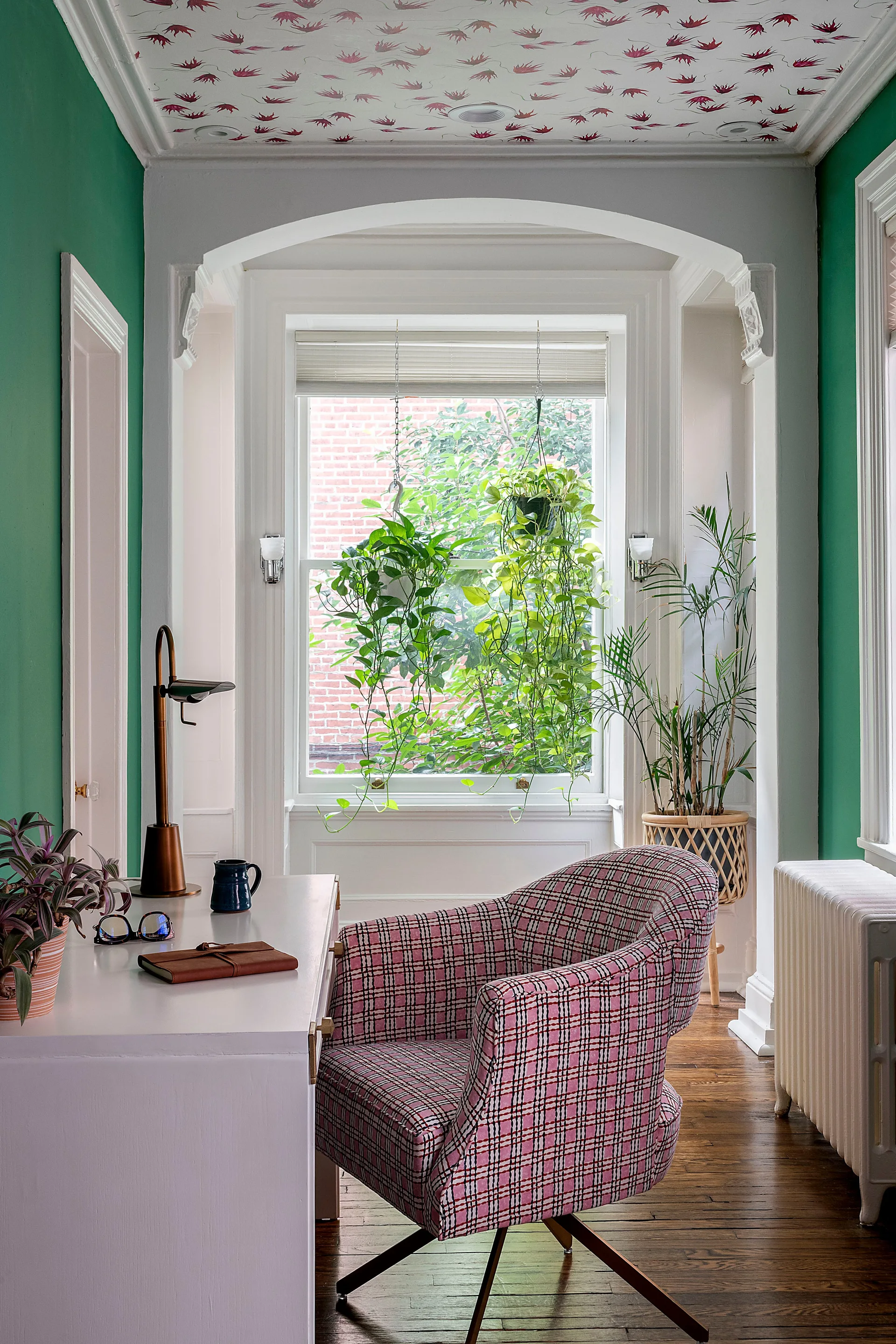 A green home office nook features a white desk, a pink plaid armchair, and a window with hanging plants under a patterned ceiling.