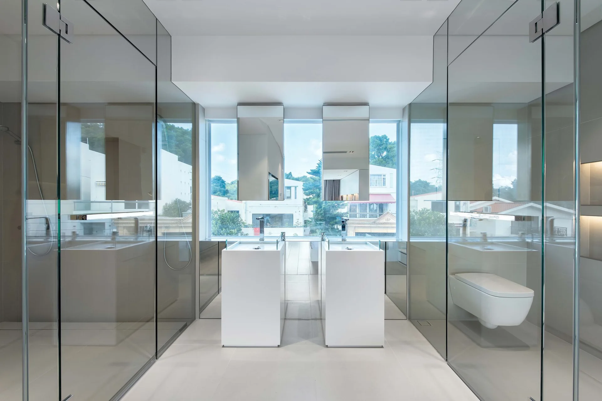 A modern white bathroom features two central pedestal sinks, rectangular mirrors hanging in front of a window, and floor-to-ceiling glass partitions.