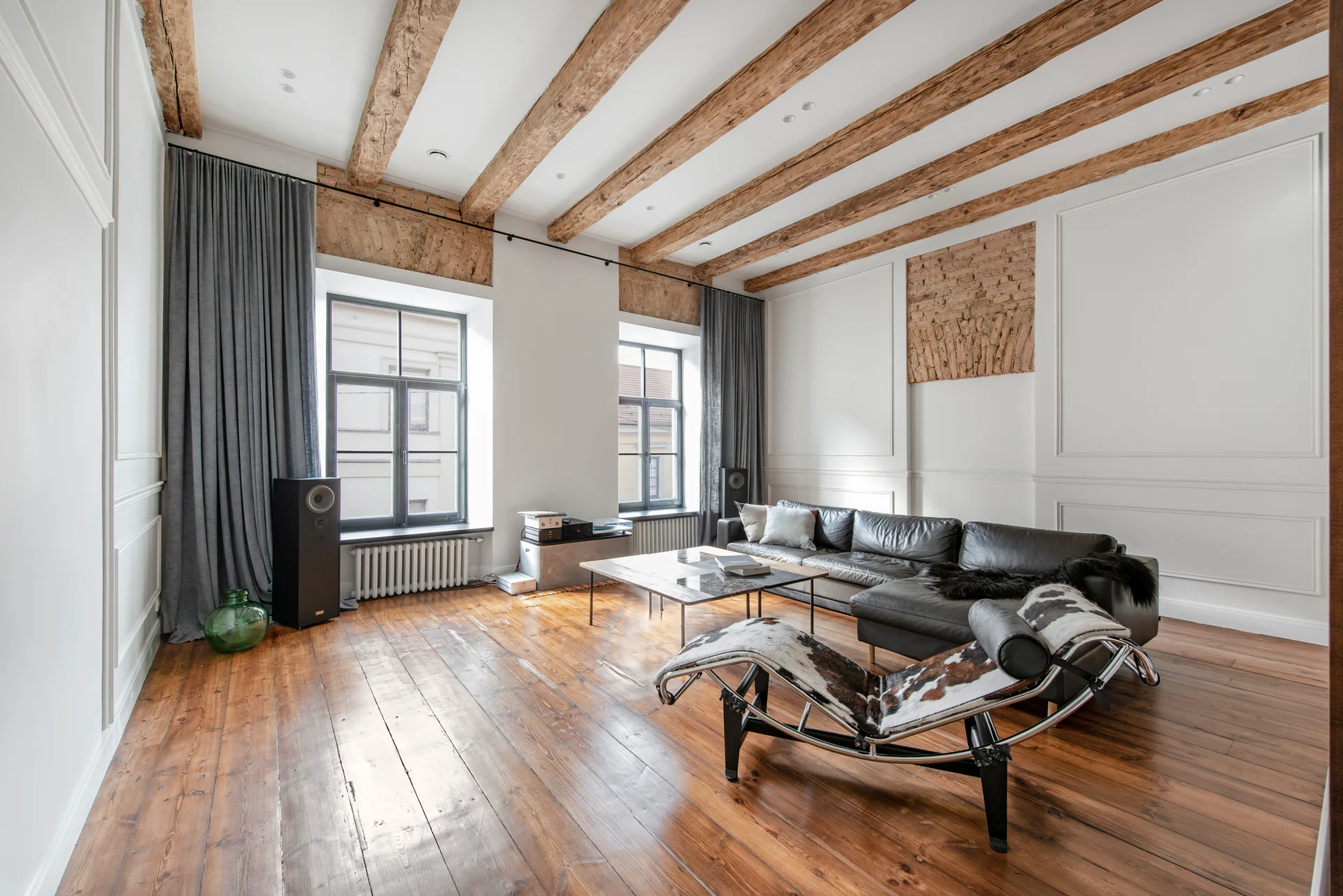 A spacious living room features exposed timber ceiling beams, white paneled walls with brick accents, grey drapes, a dark leather sofa, and a cowhide chaise.