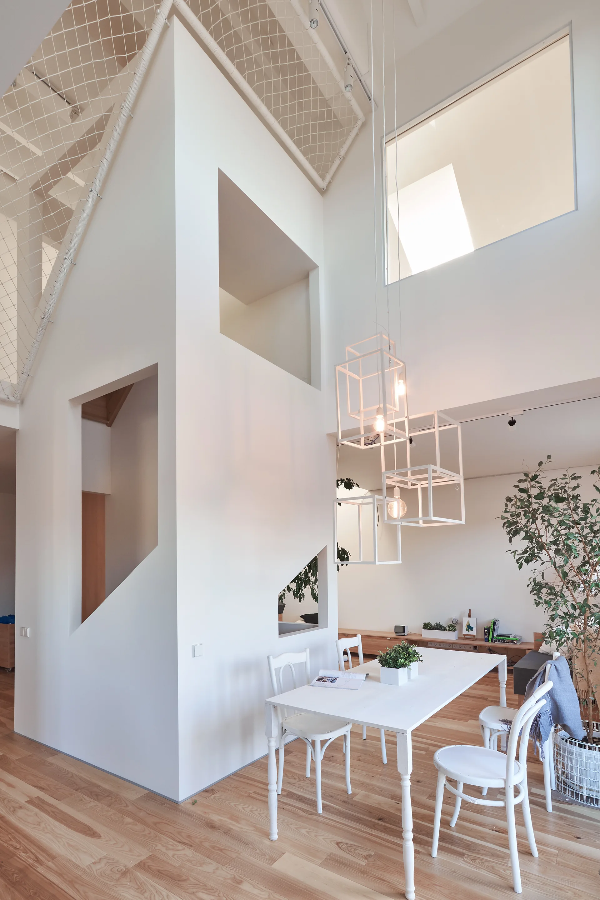 A bright minimalist dining room features a white table and chairs beneath geometric cube pendant lights next to a sculptural white staircase structure.